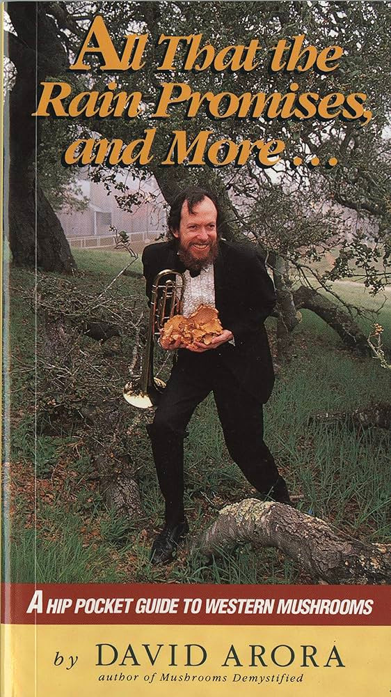 The image is the front cover of "All That the Rain Promises and More: A Hip Pocket Guide to Western Mushrooms" by David Arora. It depicts a joyful man with a beard, dressed in a formal black tuxedo, complete with a bow tie. He is standing under an overcast sky in a pastoral setting with oak trees and a wooden fence visible in the background. The man is holding a shiny brass trumpet in his left hand, and in his right, he cradles a large, fleshy cluster of golden-brown wild mushrooms. His pose suggests a spirited connection between music, nature, and the hobby of mushroom foraging.