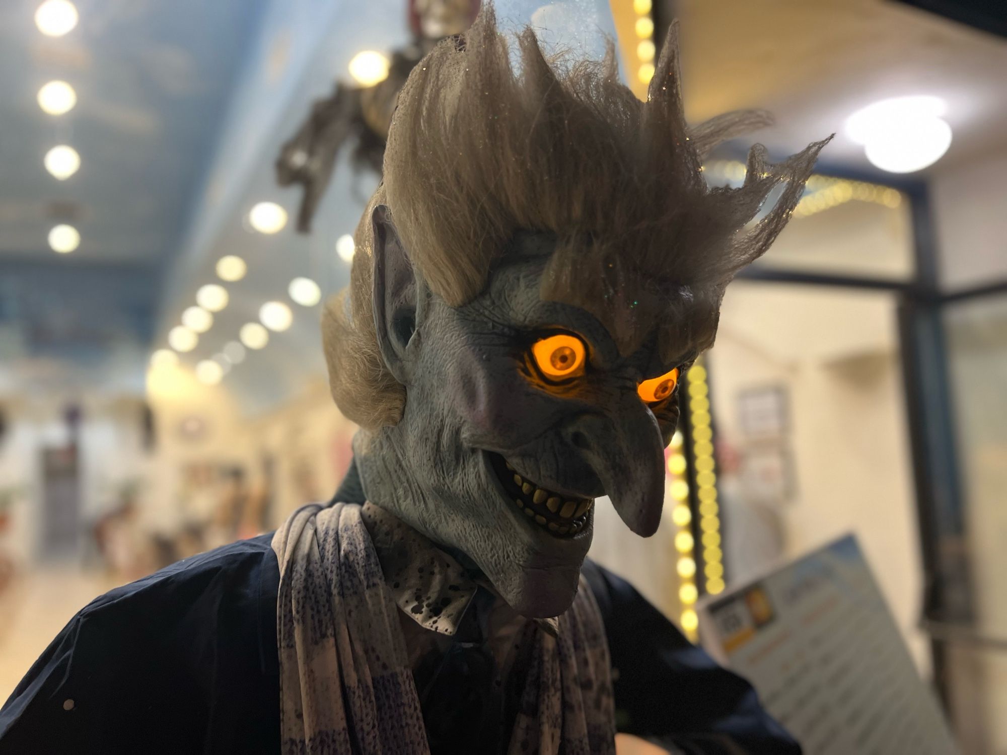 The image shows a close-up of a Halloween or horror-themed figure with glowing orange eyes. The figure has a sinister expression with exaggerated facial features, including a long, pointed nose, sharp teeth, and light-colored, wild hair. The skin is a pale blue-gray with detailed textures that give the figure a weathered or monstrous appearance. The figure is dressed in dark clothing with a patterned scarf draped around its neck. The background is softly blurred, showing a well-lit indoor area with rows of lights and some indistinct objects or people, likely part of a display or shop setting. The eerie glow from the eyes and the sharp features of the figure create a spooky atmosphere.