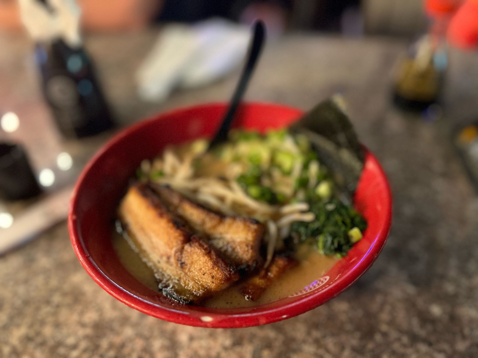 The image shows a close-up of a bowl of ramen in a red bowl, placed on a table. The ramen features two slices of grilled pork belly, green onions, leafy greens, bean sprouts, and a sheet of nori (seaweed) on the side. The broth is light in color, likely a tonkotsu or miso base. A black spoon is placed in the bowl, ready for use. The background is softly blurred, giving focus to the bowl of ramen, with some out-of-focus items like a napkin and other condiments visible on the table. The overall scene suggests a warm and inviting meal.