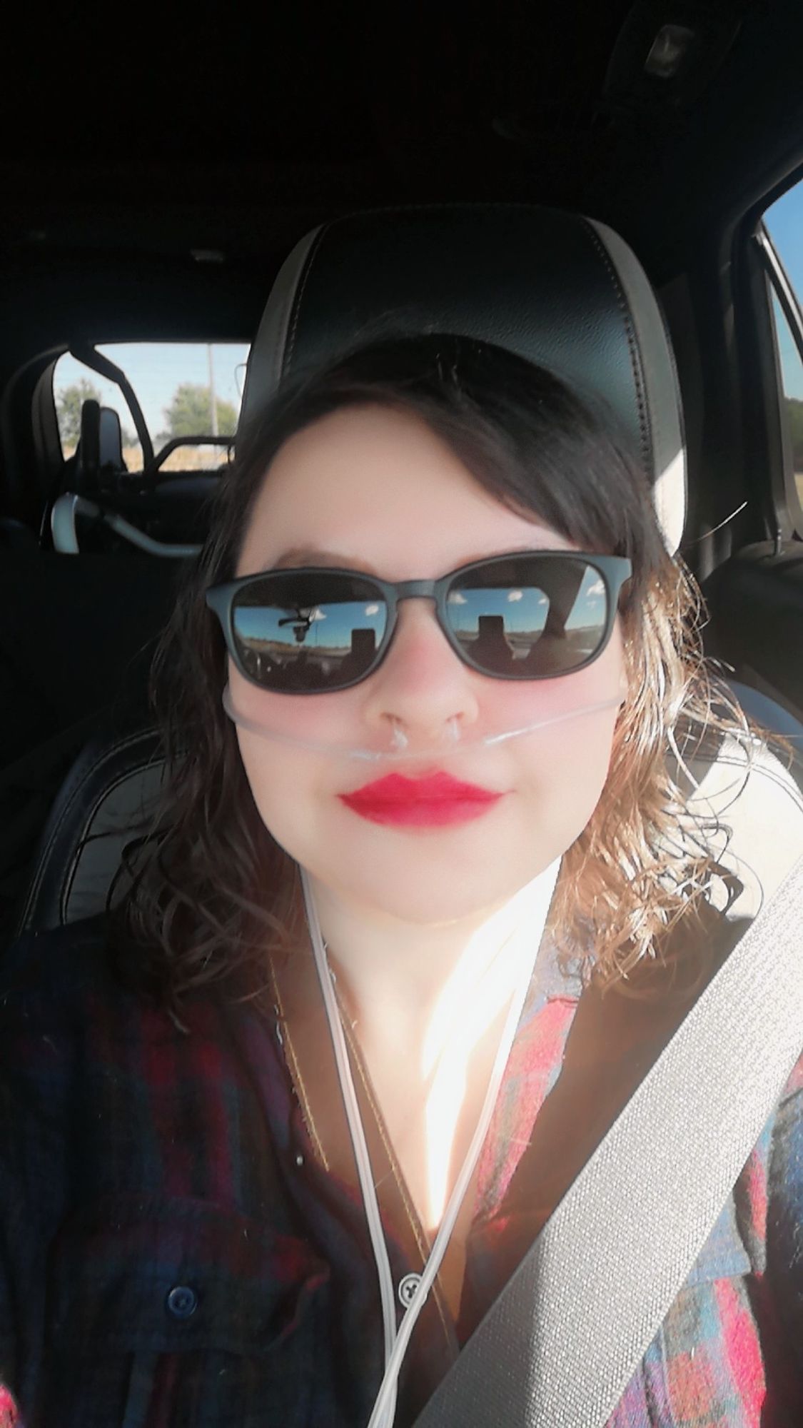 me in the passenger of the car wearing black sunglasses, a purple, red and blue plaid button down top, red lipstick and my oxygen tubes.