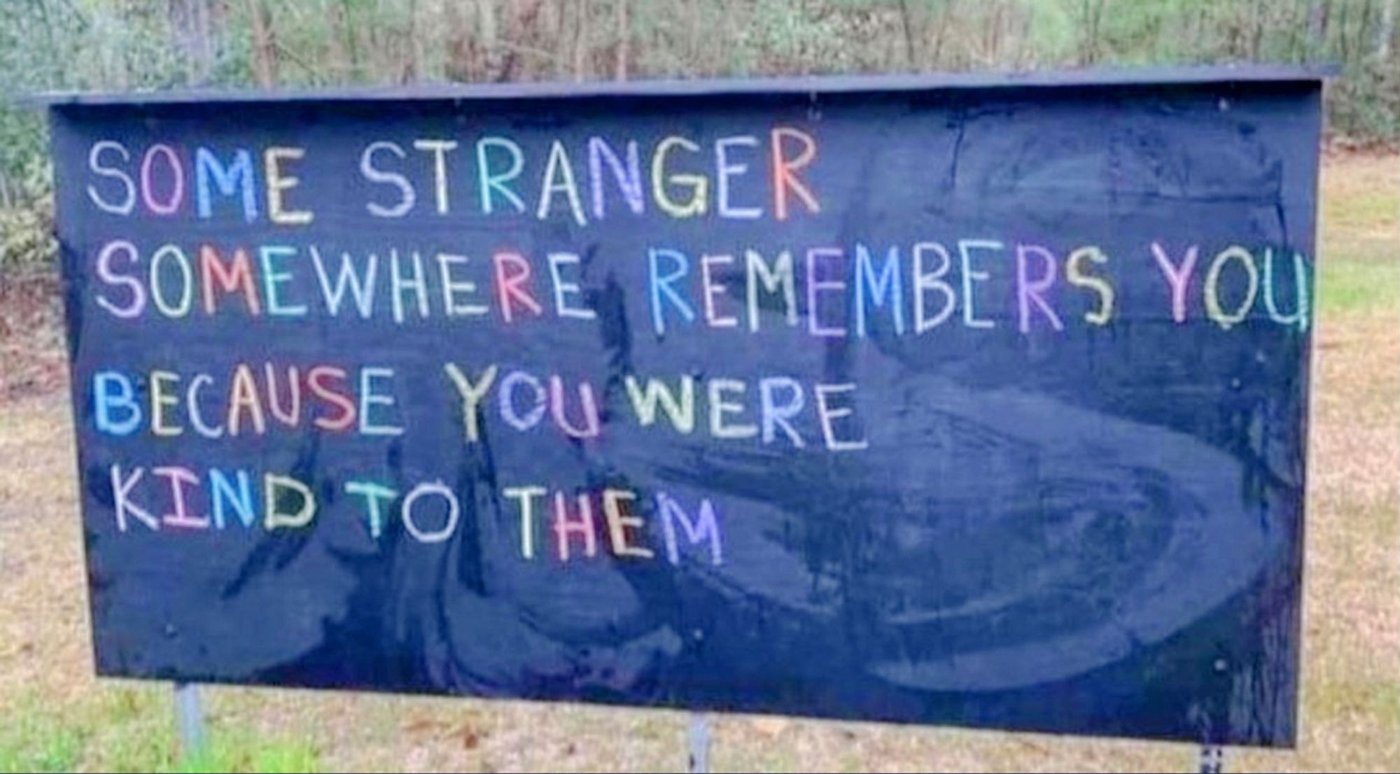 Posted outdoors on a clear day on a grassy area, surrounded by green trees, is a rectangular blue banner, with these words written on it in colorful letters:
Some stranger, somewhere,
remembers you, because
you were kind to them.