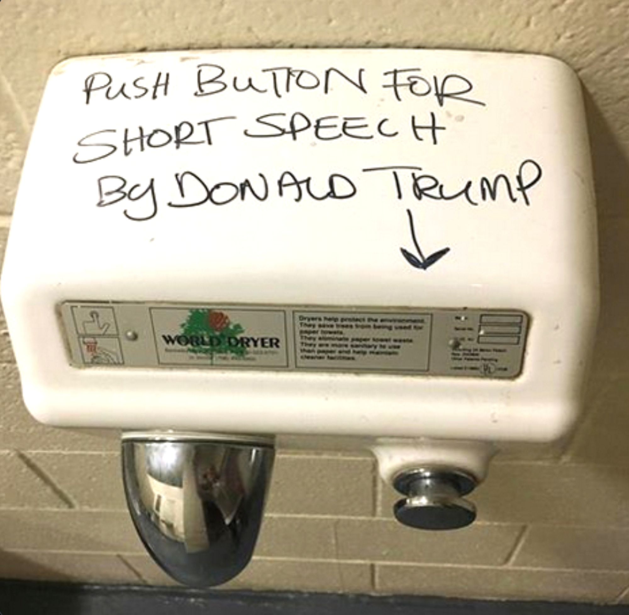 A white, mechanical hand drying machine with a silver-colored blower nozzle and push button affixed to a public restroom wall. Written on its top is, "Push button for short speech by Donald Trump."