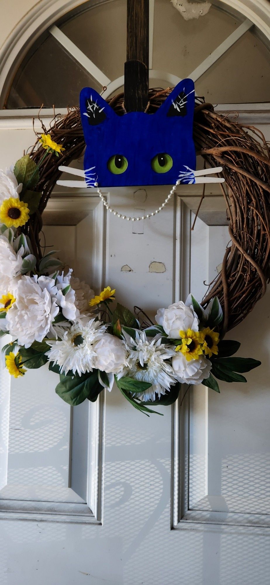 A door wreath with white flowers and small sunflowers, with a blue cat half-face, that has white whiskers, green eyes and a pearl necklace.