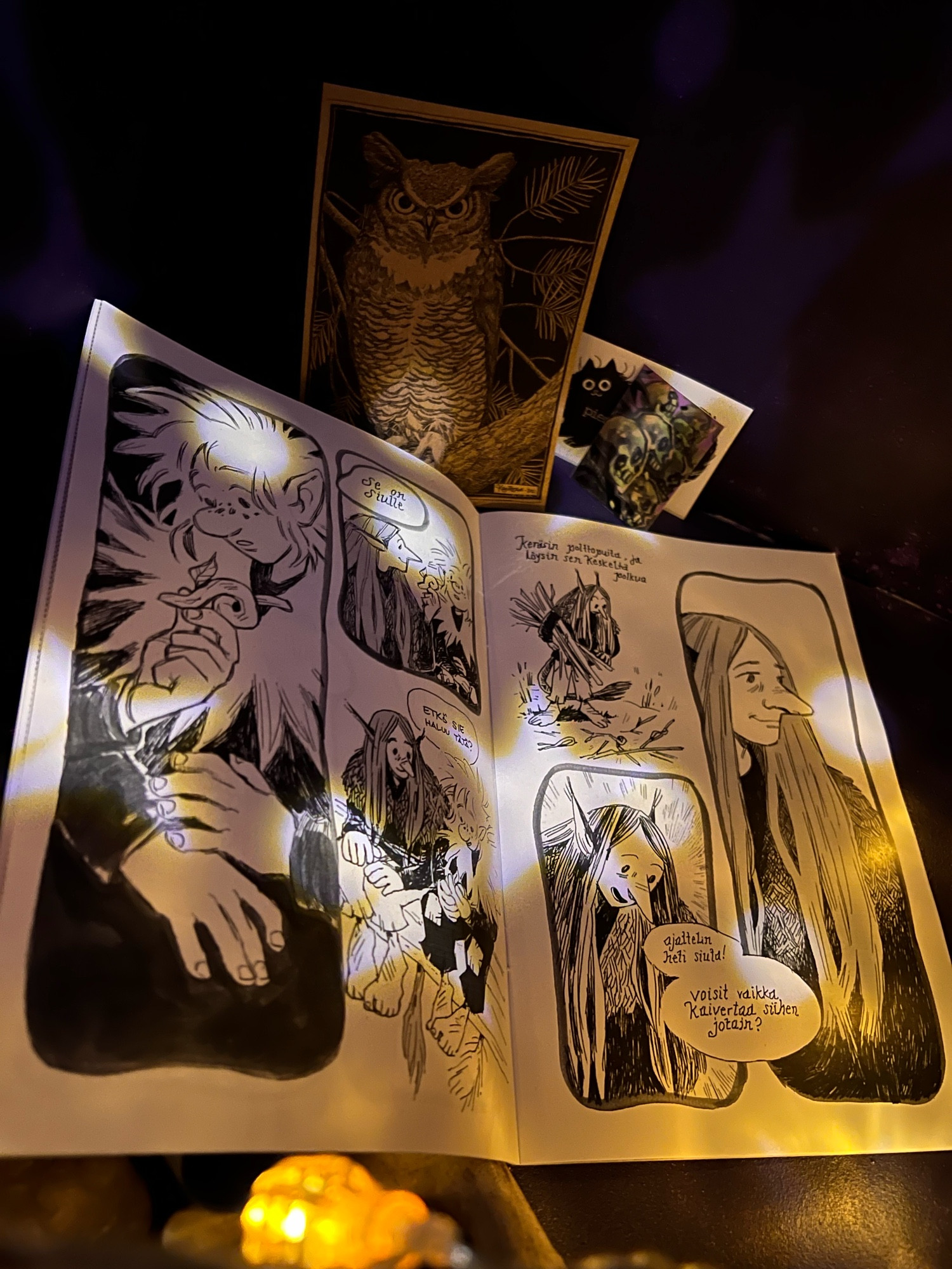 An open spread from the comic zine Kamu by the artist Käenretale, which features several panels of of two troll friends inked in black and white. In the background there is a small print of an owl, a sticker of a black cat and another with several skulls on it. 

The comic is in Finnish (English in brackets below) and shows Oksa, a fluffy haired troll, receiving a curled stick from Nuppu, a troll with long straight hair.
Nuppu: “Se on siulle” (It’s for you)
Oksa: ”Etkö sie haluu tätä?” (Don’t you want it?)
Nuppu: ”Keräsin polttopuita, ja löysin sen keskeltä polkua. Ajattelin heti siuta! Voisit vaikka kaivertaa siihen jotain?” (I was collecting firewood and found it in the middle of the path. I thought of you right away! You could carve something into it?)