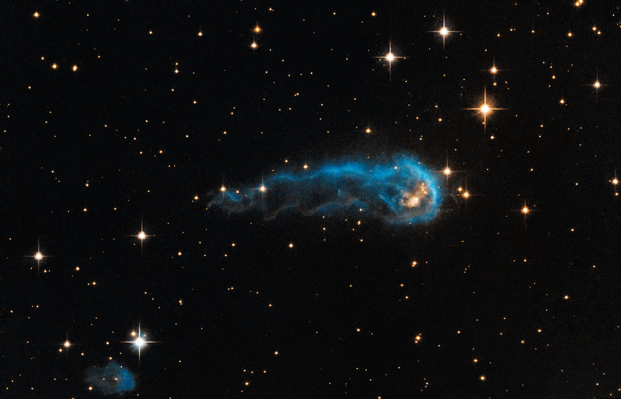 A forming protostar with an envelope of gas around it. To the right of center, there is a glowing orange knot of gas, the protostar, surrounded by a dark blue cloud of dust and gas. The blue dust and gas trails into a comet-like tail that points to the left. White and yellow stars are spread throughout the image.