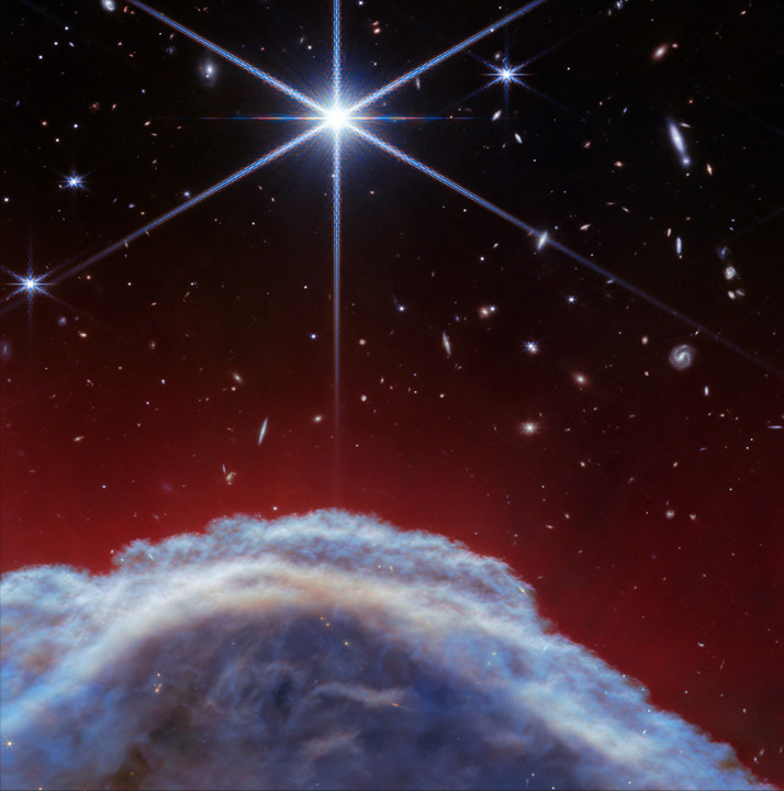 A clumpy dome of blueish-gray clouds rises about a third of the way from the bottom. Above it, streaky, translucent red wisps brush upward to about halfway up the image. The top half of the image is the black background of space with one prominent, bright white star with Webb’s 8-point diffraction spikes. Additional stars and galaxies are scattered throughout the image, although very few are seen through the thick clouds at bottom and all are significantly smaller than the largest star.