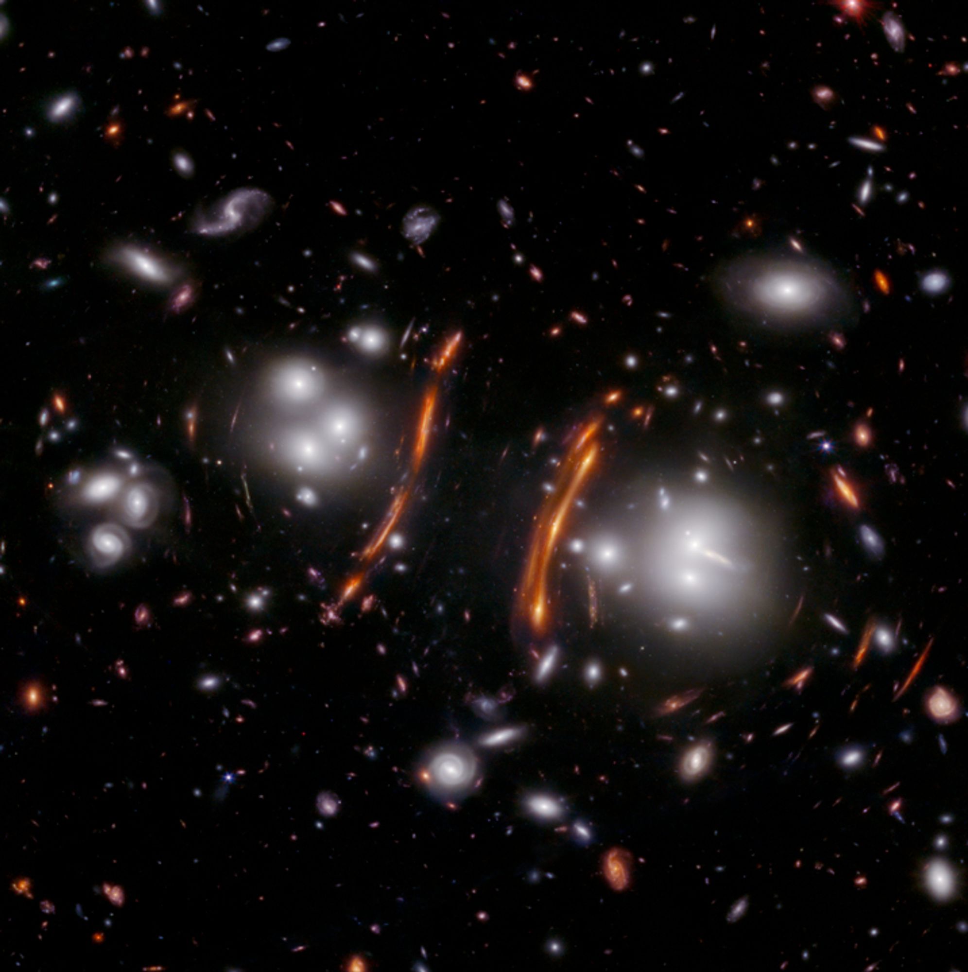 A black background is scattered with hundreds of small galaxies of different shapes, ranging in color from white to yellow to orange to red. Some galaxies are distorted, appearing to be stretched out or mirror imaged. Just to the left of the center of the image, there is a long, red arc that stretches from 2 o’clock to 7 o’clock. At its left, is a cluster of a few white galaxies that look like a glowing orb. To the right of the center of the image, the red arc and glowing orb of galaxies at the left appear to be mirrored. The right-hand red arc stretches from 3 o’clock to 5 o’clock, the glowing orb of galaxies is at its right.