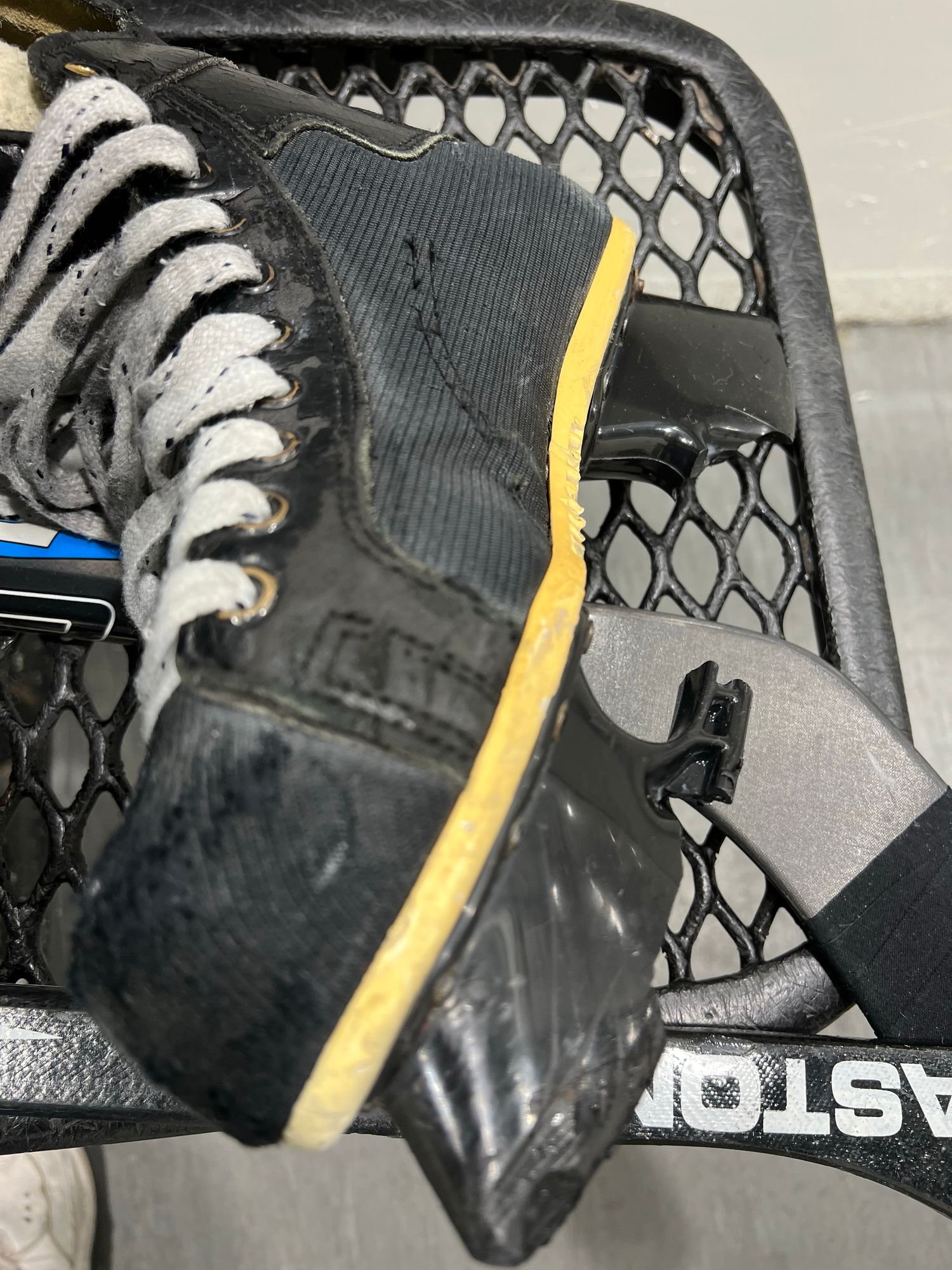 A close-up image of a black hockey skate resting on a black mesh surface. The skate  blade and structure are shattered and mostly gone