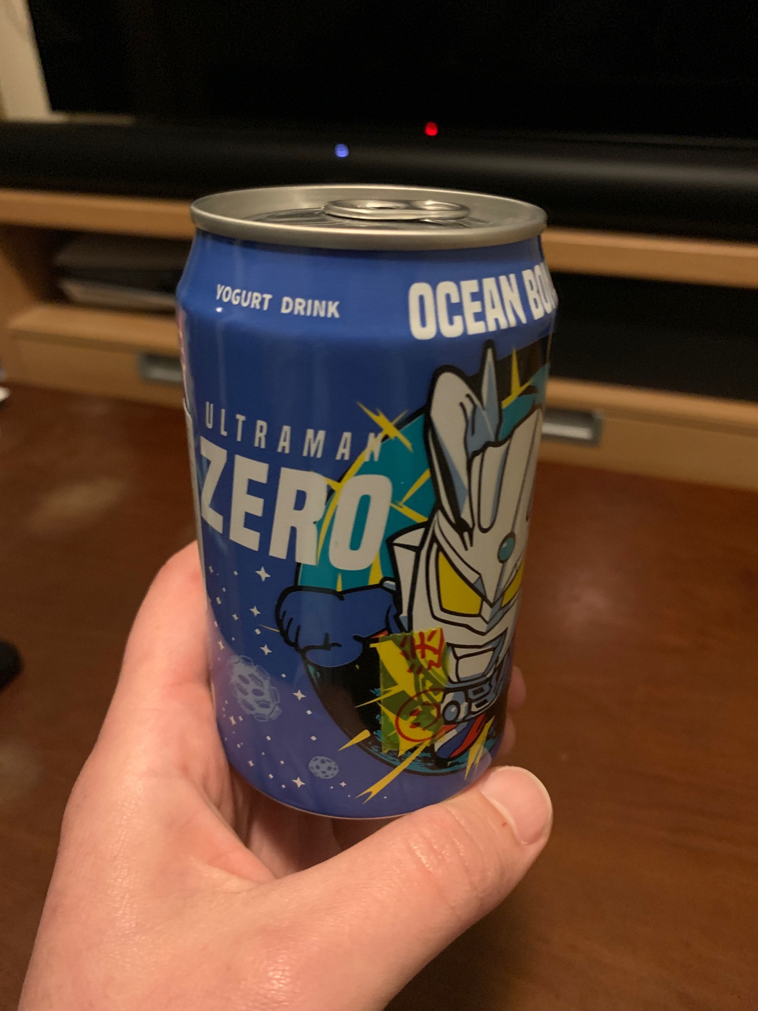 A hand holding up a blue canned beverage called Ultraman Zero. It's "yoghurt drink" flavoured, with Ocean Bomb branded along the top. A stylized illustration of the Japanese character Ultraman is emblazoned across the front, punching the air.