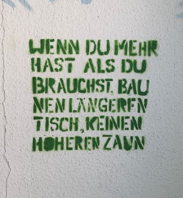 Auf eine weißgraue Wand steht mit grünen Großbuchstaben gesprüht:
WENN DU MEHR HAST ALS DU BRAUCHST,  BAU NEN LÄNGEREN TISCH, KEINEN HÖHEREN ZAUN