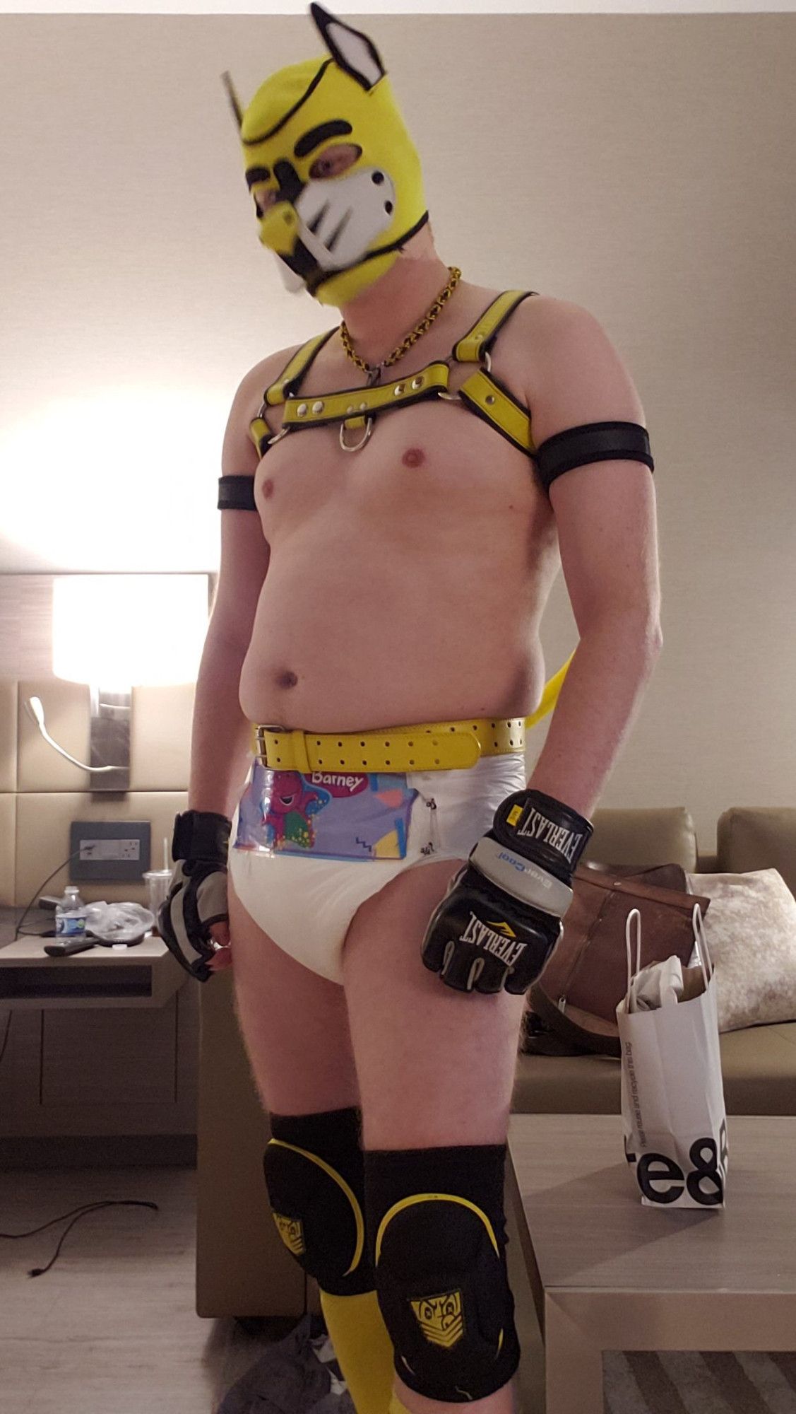 A man in a pup hood, leather harness and diaper standing upright showing off his outfit.