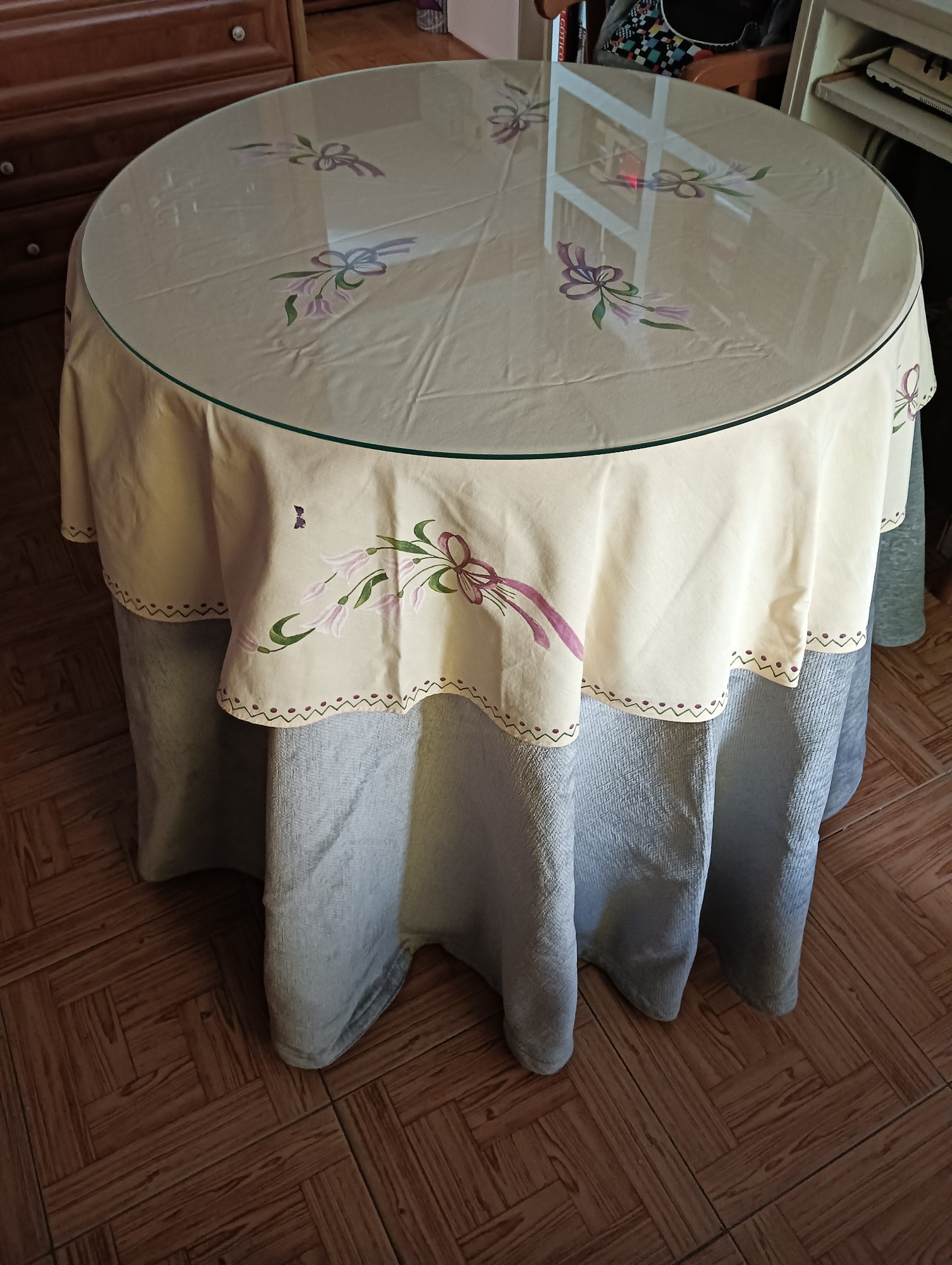 Una mesa camilla con faldilla gris y tapete de color crudo con calas blancas y rosas pintadas.