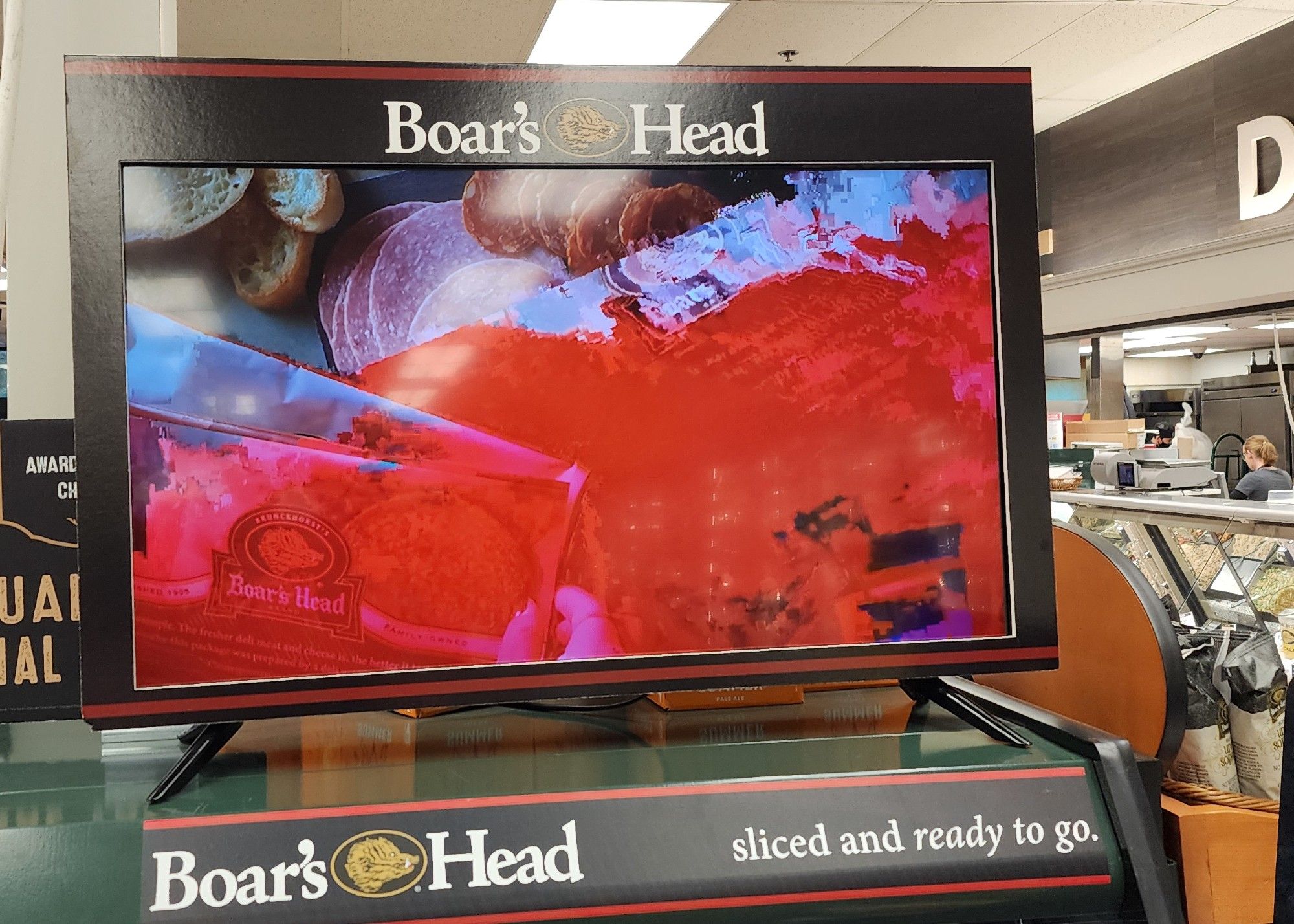 A photo of a grocery store kiosk for Boar's Head Meats. On a monitor, a smeared red-and-cyan decoding artifact obscures most of the image of a platter of meat and bread.