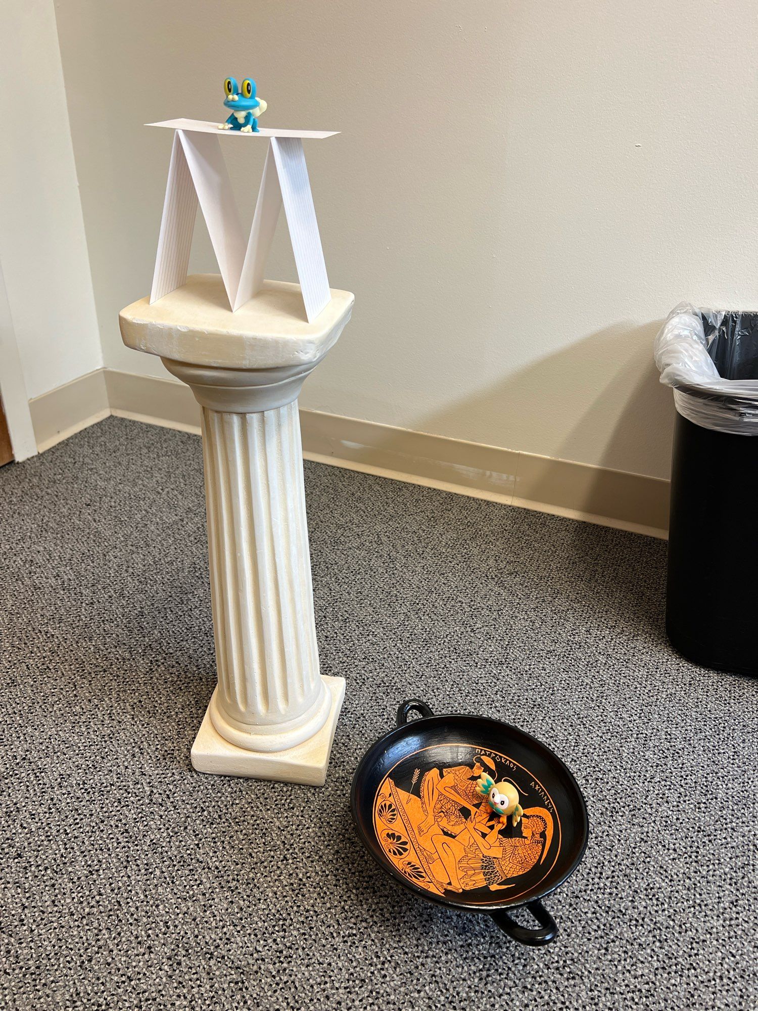 A small Doric pillar replica with an index card tent and a Froakie figurine on top to the left, a replica of a Greek kylix with a Rowlet figurine inside to the right