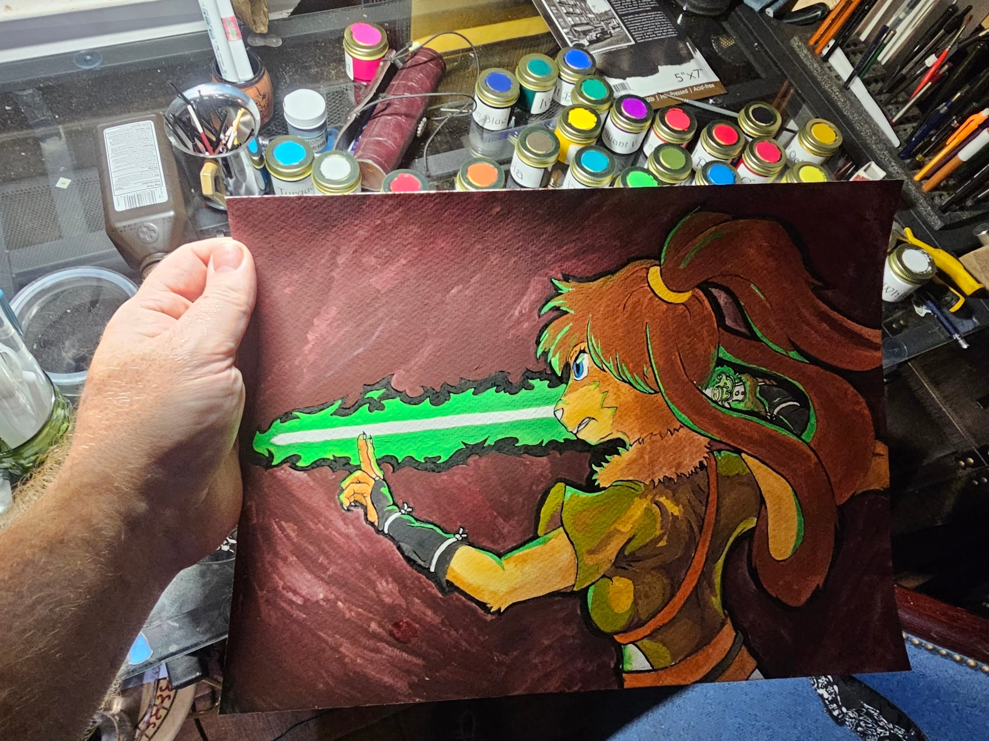 A hand holding a painting of Lop, a character from star wars visions holds her lightsaber in a way that slightly mimics a traditional Tai chi sword pose. A desk in the background has jars of paint on it.