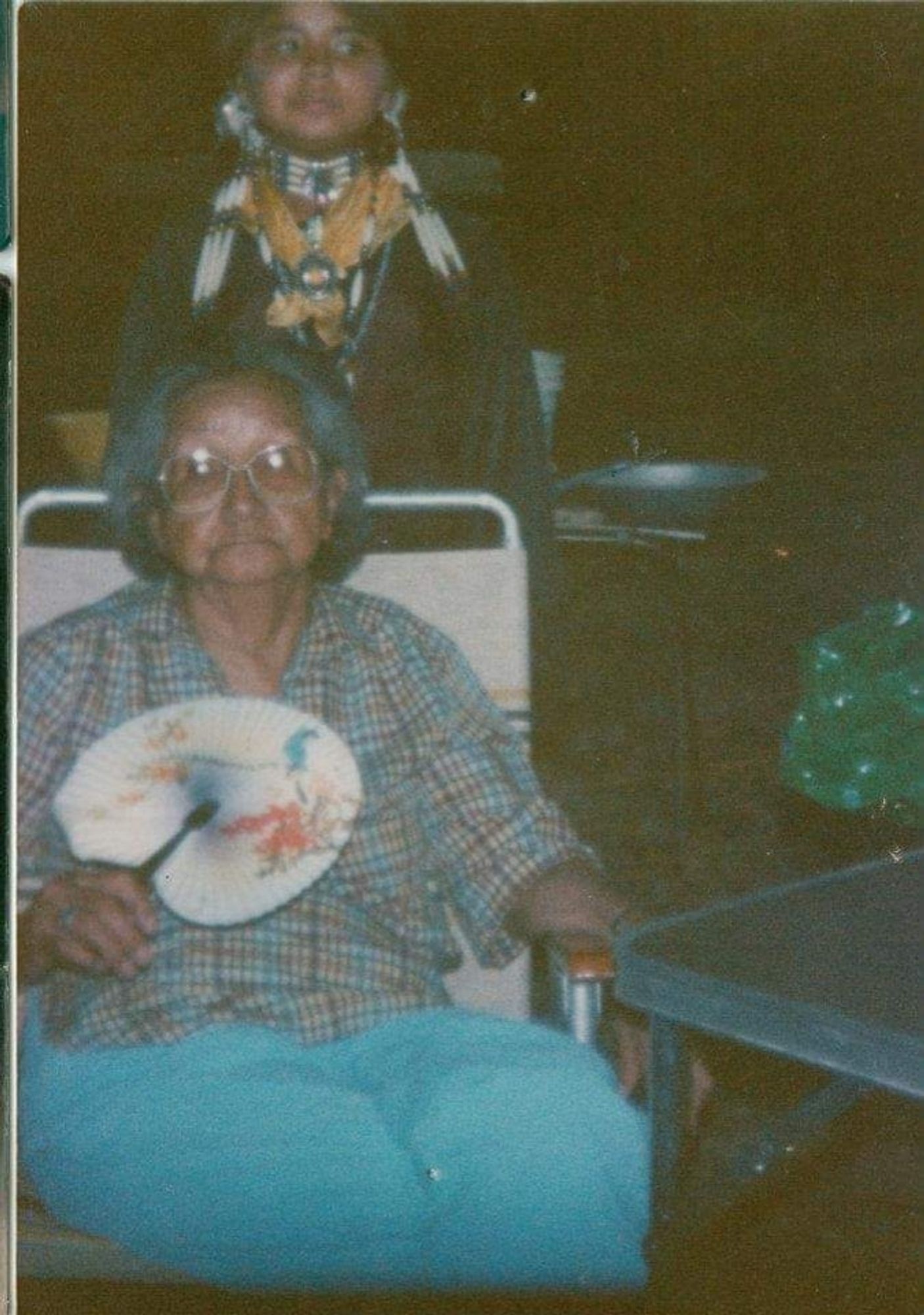 A photo of Johnnie's great grandma, Minnie Moore wearing turquoise pants and a plaid shirt sitting on a chair while holding a white handheld fan. Behind her stands a young Johnnie Jae in her Native regalia.