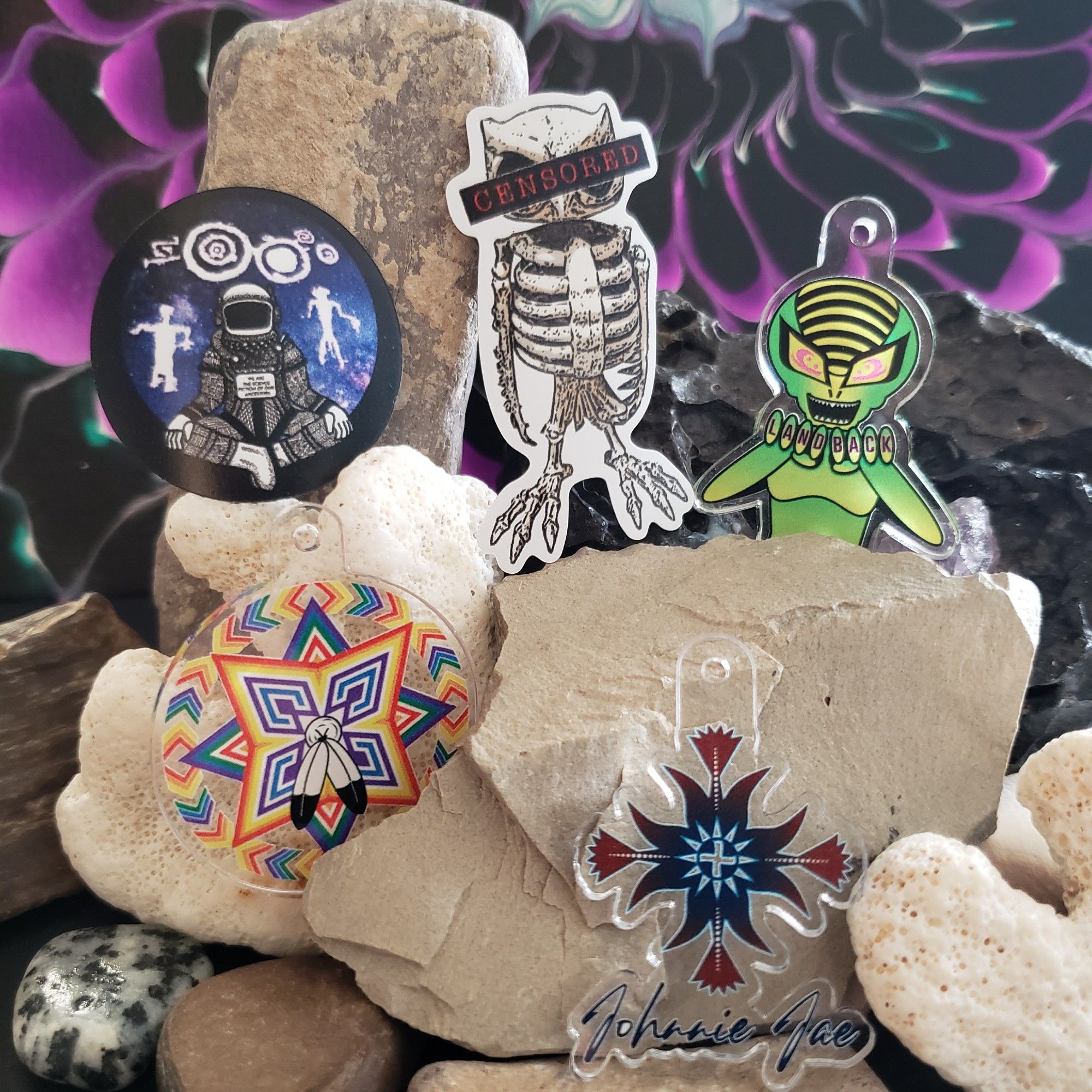 A Collection of magnets and acrylic charms posed against white rocks. From Left to Right (top row): A round 1.5in magnet featuring an astronaut surrounded by Native American rock drawings, a 1.5in magnet of a skeletal owl with a censored banner across its eyes, an acrylic charm of a bright neon green alien with Land Back written across her knuckles. From Left to Right (bottom row): An acrylic charm featuring a geometric design inspired by the Two-Spirit Pride flag, an acrylic charm featuring the Legacy Floral Logo.