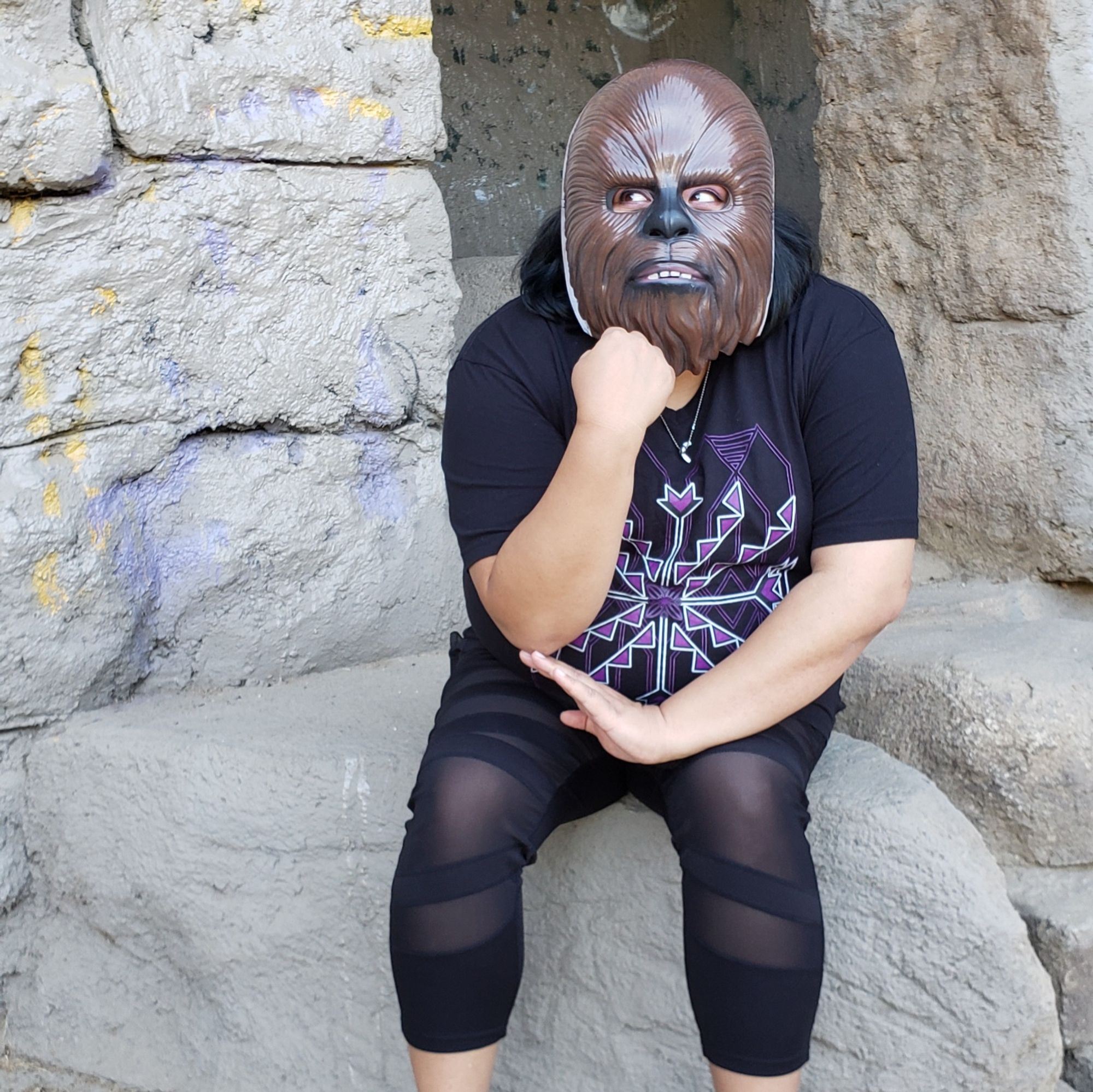 Johnnie Jae sitting on a rock like the Thinker, her hand under her chin while wearing a wookie mask and giving major side eye.