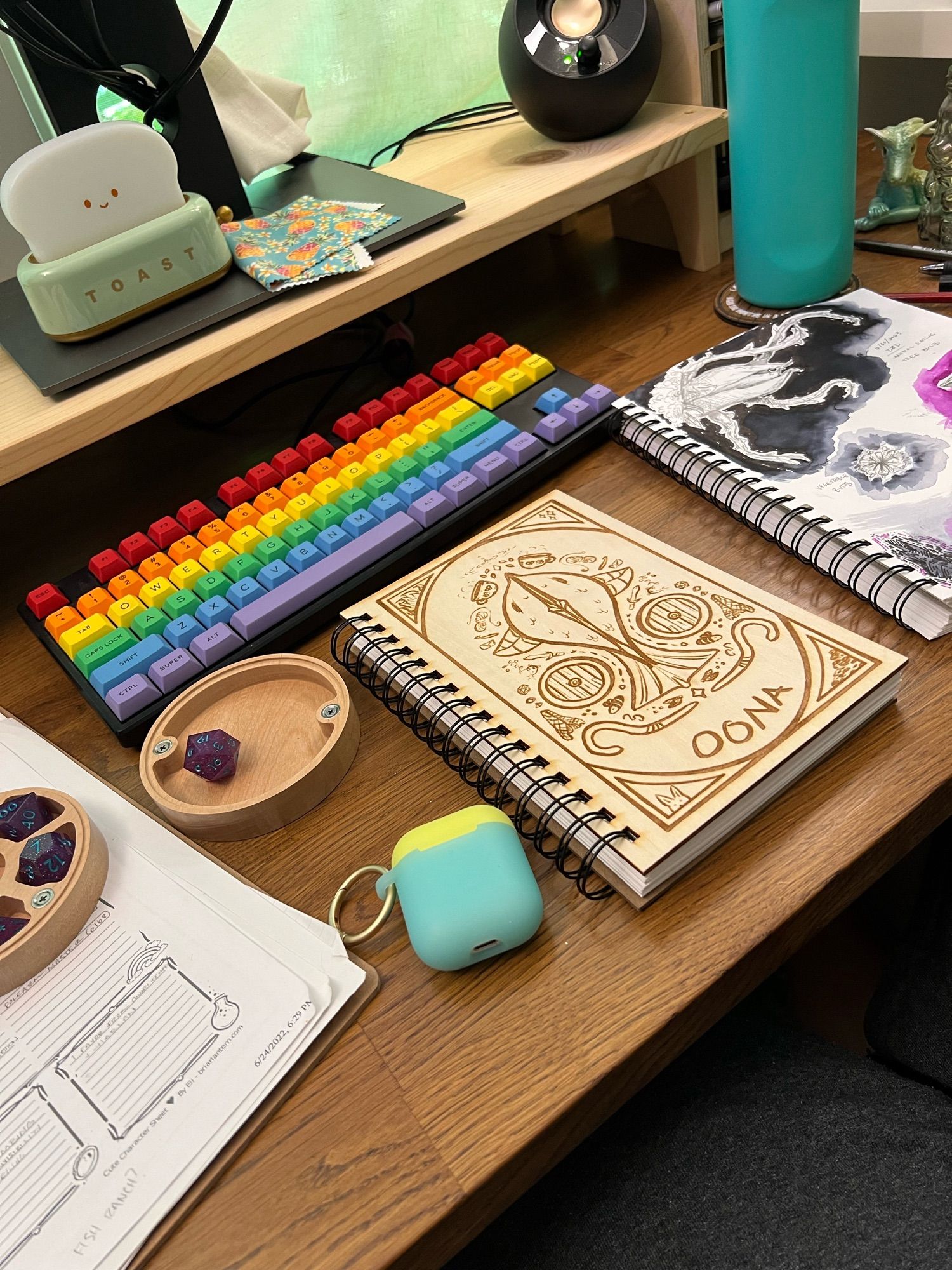 Wooden desk with various items (including notebooks, sketchbooks, papers, polyhedral dice, etc.) with a custom, laser engraved notebook in the center with d&d character themed items on the cover.