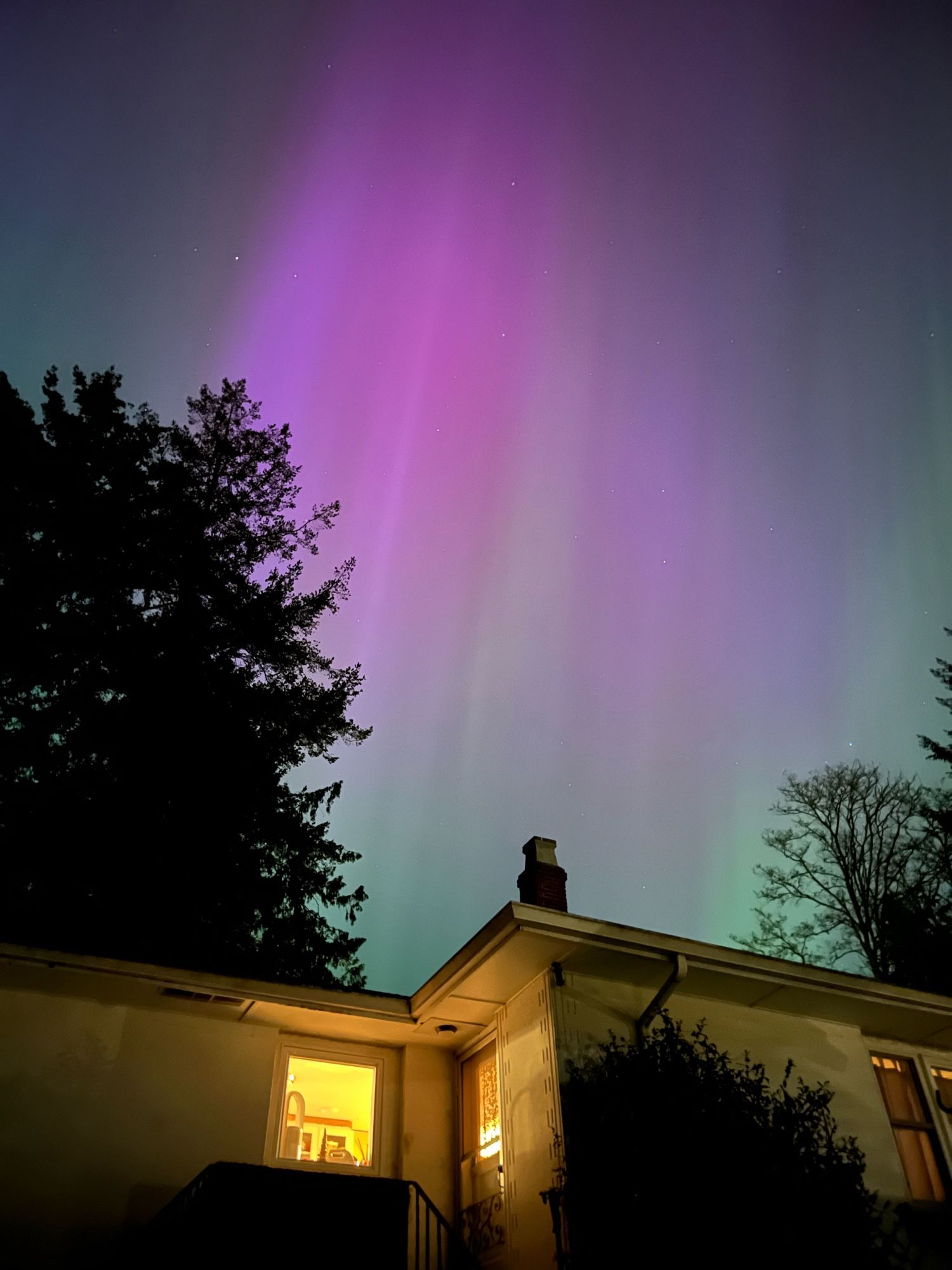 Purple and green northern lights in a sky with a few scattered stars with the exterior of a house in the foreground glowing yellow from the lights emanating from inside