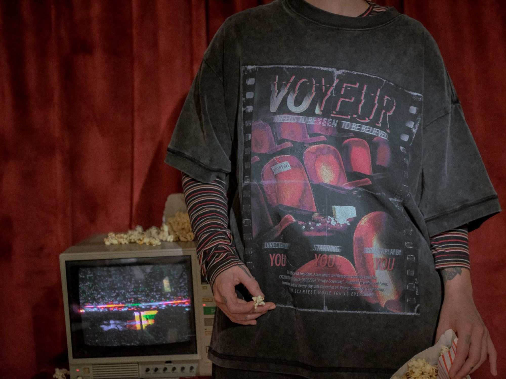 A person modelling The Magnus Protocol x Knock Thrice merchandise. They are standing in front of an old, glitched screen, and holding popcorn. They are wearing a black, stone-washed t-shirt with a cinema-inspired design and the title "Voyeur".