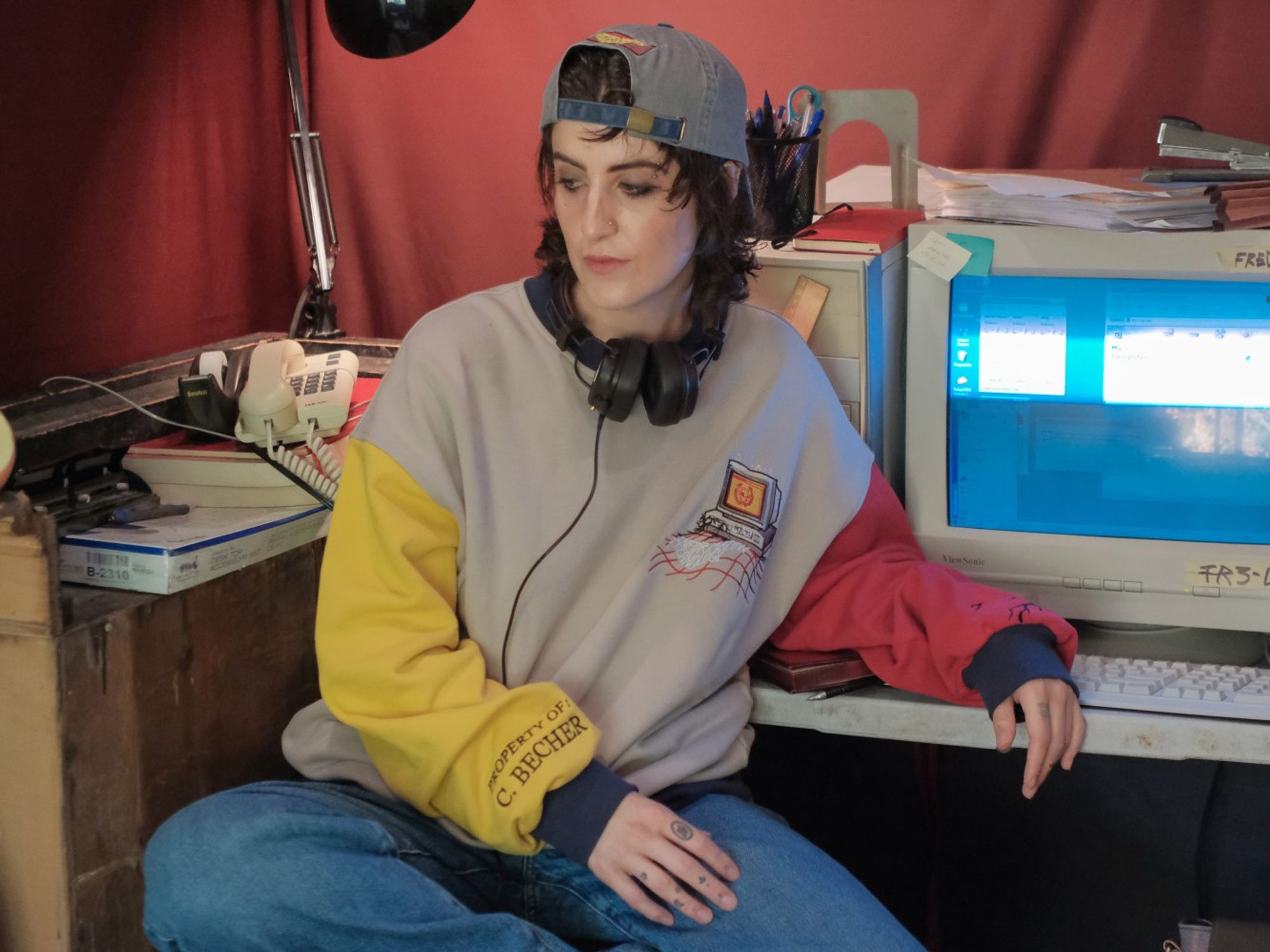 A person modelling The Magnus Protocol x Knock Thrice merchandise. They are sitting in front of an old computer, with a glitched broken screen. They are wearing a baseball hat back to front. The cap is grey and features embroidering reading "information technology". They are also wearing a colour-blocked crewneck. The body of the crewneck is grey, the right arm is red and the left arm is yellow. The cuffs are navy blue. The crewneck features an embroidered computer, and on the sleeve is embroidered "Property of C. Becher".