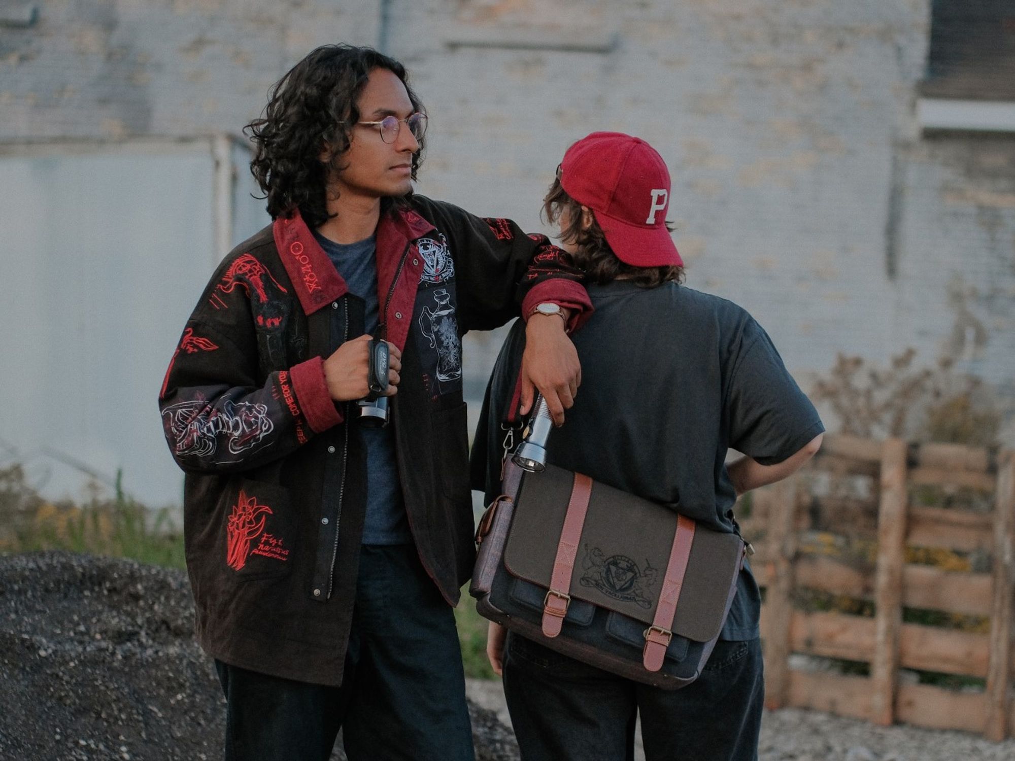 Two people model The Magnus Protocol x Knock Thrice merchandise. The person on the right has their back to the camera is wearing a messenger bag featuring a logo for the OIAR from The Magnus Protocol. The person on the left is facing the camera, but looking off to the right. They are wearing a black denim jacket with red accents. The jacket is decorated with numerous patches and embroidery, inspired by alchemy.