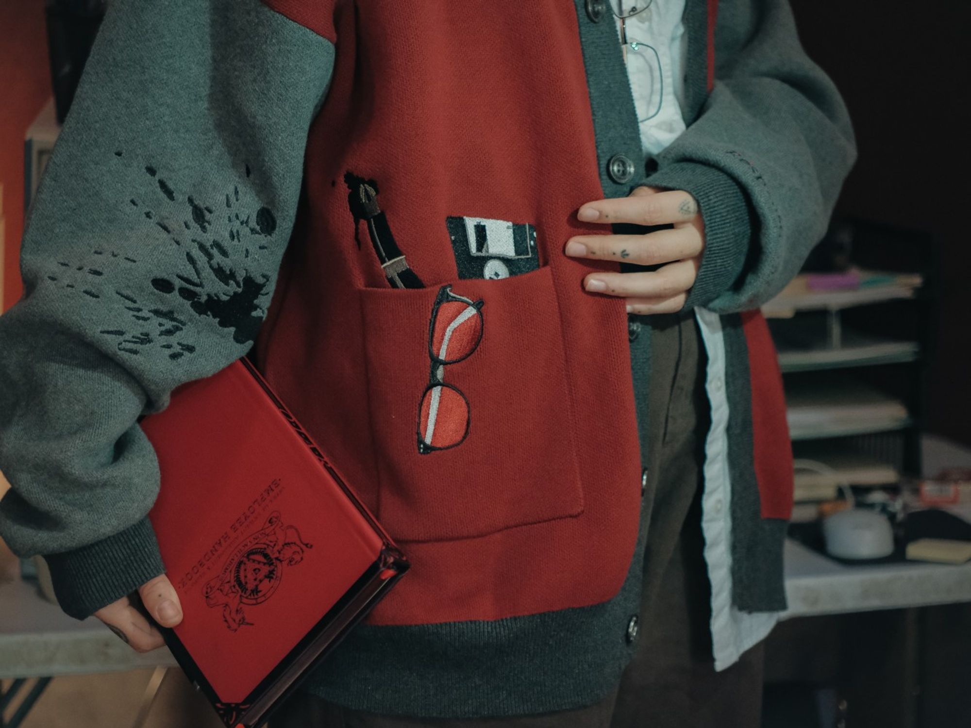 A person modelling The Magnus Protocol x Knock Thrice merchandise. This photo is of the detail of a red and grey cardigan. The arms of the cardigan are grey, and the body is dark red. Embroidered on the cardigan is a pen, floppy disc and pair of glasses, made to seem as if they're coming out of a pocket. On the sleeve is an embroidered ink stain. The model is also holding a faux-leather red journal, featuring the OIAR logo and the words "Employee Handbook".