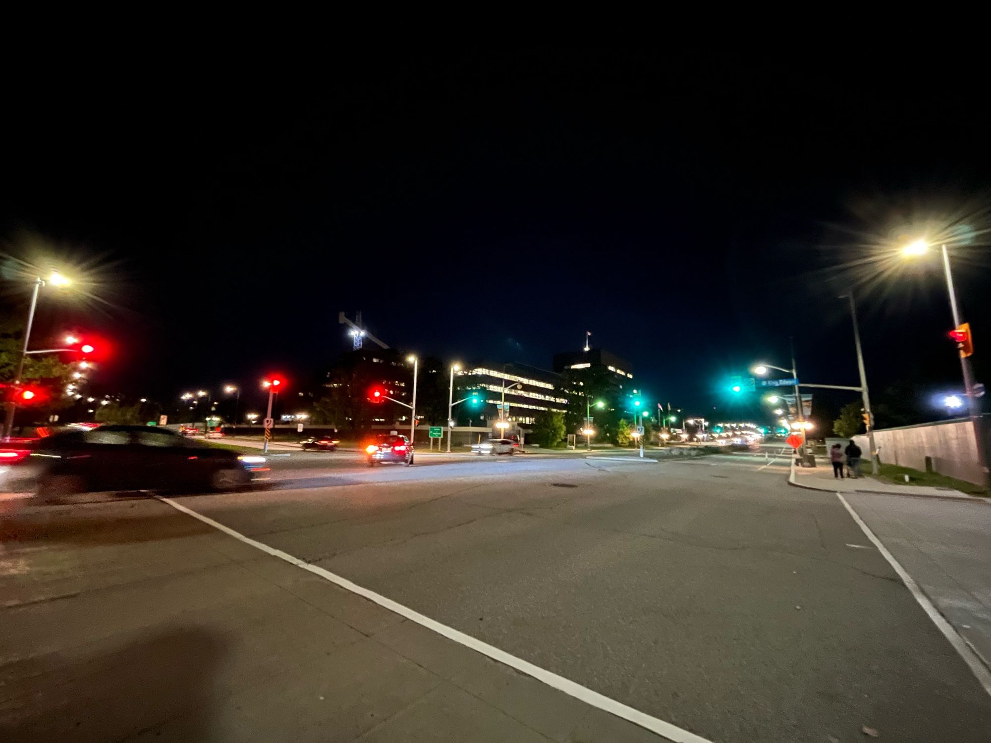 Une intersection la nuit. On voit beaucoup d’asphalte au premier plan, quelques voitures floues, des rouges à gauche et vert à droite, et un bâtiment éclairé en arrière-plan. 
An intersection at night. There’s lots of asphalt in the foreground, a few blurry cars, red lights on the left and green lights on the right, and a building in the background.