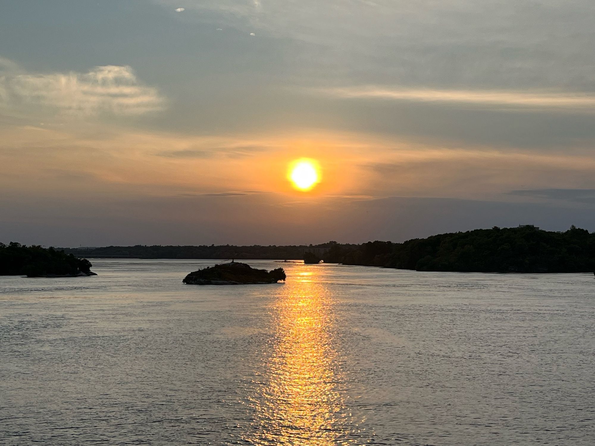 Photo d’une île au milieu de la rivière des Outaouais au coucher du soleil. Le soleil arrive presque à la ligne d’horizon. Le reflet du soleil s’étire jusqu’en bas de la photo. Le ciel est en partie voilé et va du gris au-dessus des arbres au orange autour du soleil et au bleu gris en haut.