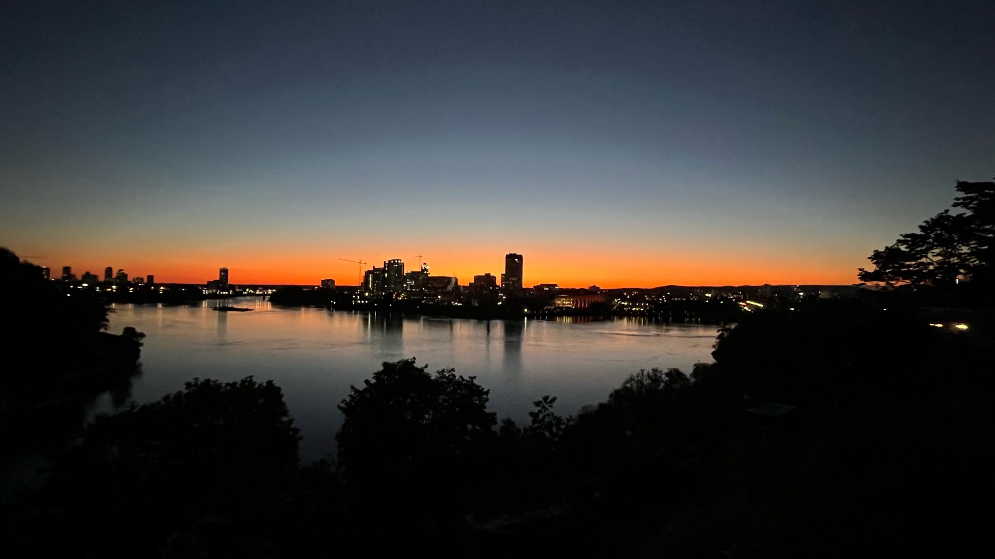 Vue de Hull au coucher du soleil. Au premier plan, les buissons et arbres forment une masse noir dont on voit le contour. Au milieu, comme un lac entouré d’arbres, la rivière des Outaouais. Le pont du Portage et la silhouette de Hull coupent l’image en deux. Au-dessus, le ciel qui va de l’orange au bleu gris clair et au gris foncé en haut.