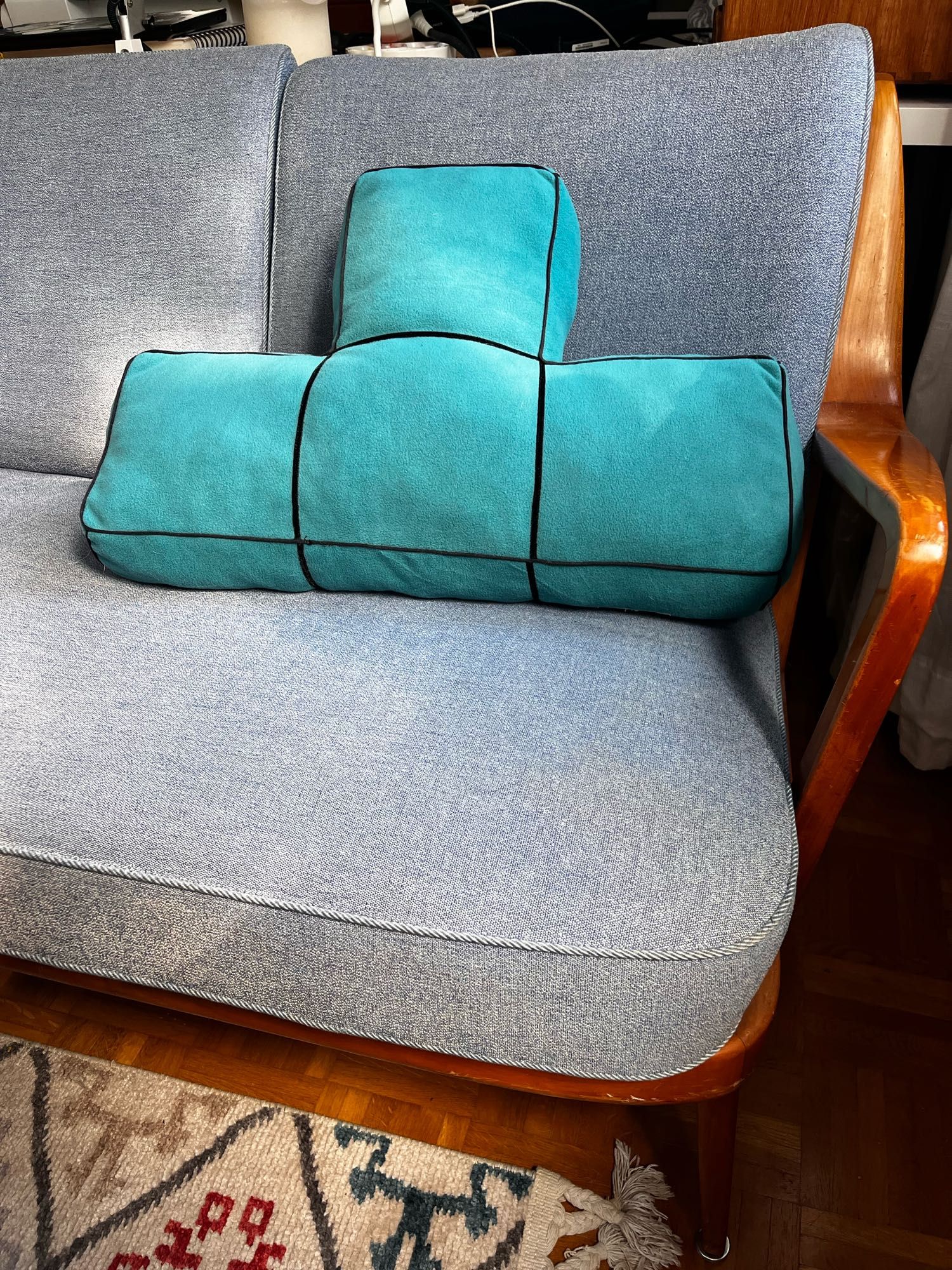 Auf einem blauen Sofa liegt ein Petrol farbenes Kissen in Form eines T. Wie bei Tetris üblich hat das Kissen einen schwarzen Rahmen durch schwarzes Paspelband das in der Außennaht eingefasst ist. Es hat außerdem eine Einteilung in die vier Blöcke durch schmales schwarzes Samt Band, das auf den Stoff aufgesteppt ist.