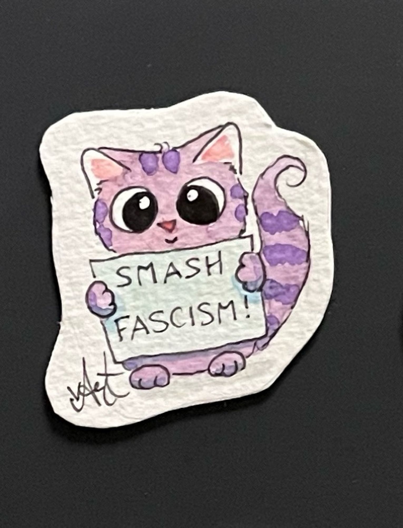 Kleines Aquarellbild: eine rosa Katze mit lila Fellmuster und buschigem Schwanz schaut mit großen runden Augen. In den Vorderpfoten hält das Kätzchen ein Schild auf dem steht: „Smash Fascism“.