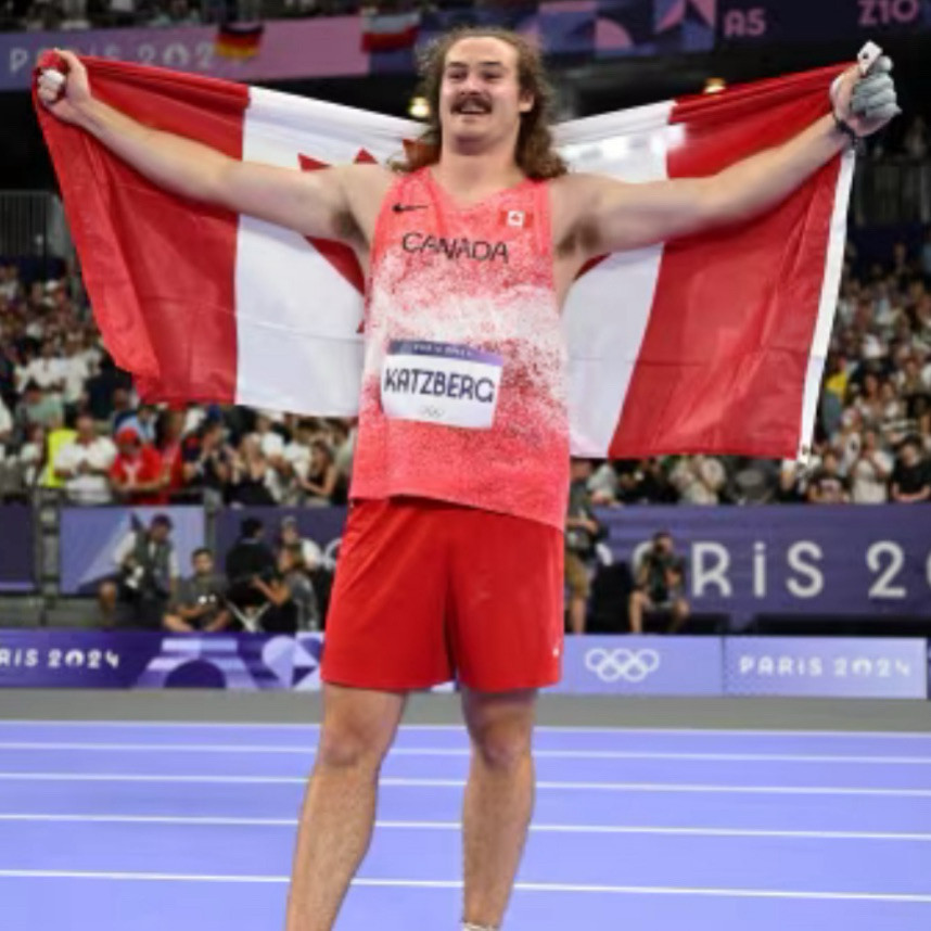 A person with long brown hair and a moustache wearing athletic gear and holding up a Canadian flag.