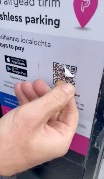 A close-up of a hand peeling off a QR code sticker that partially covers another QR code on a parking payment sign. The sign has text in both English and Irish, indicating “Cashless parking” and providing ways to pay through mobile apps.