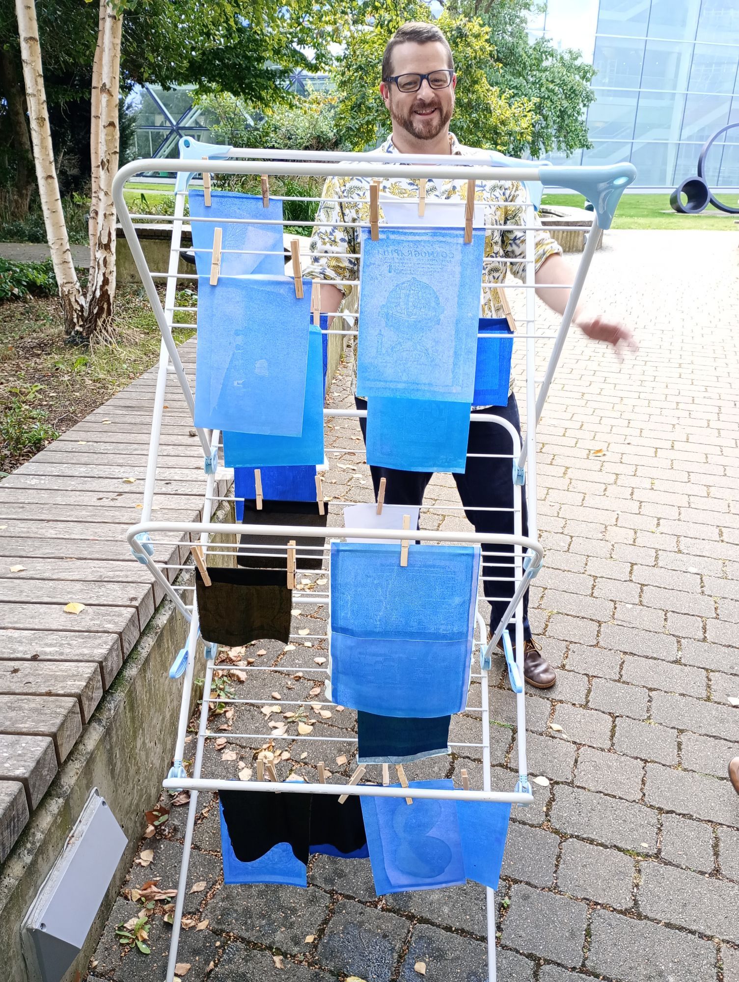 Cyanotypes drying in the sun: A clothes rail with multiple pieces of blue paper and fabric - some with visible patterns or text - pegged to it. A man in a yellow floral shirt stands in the background