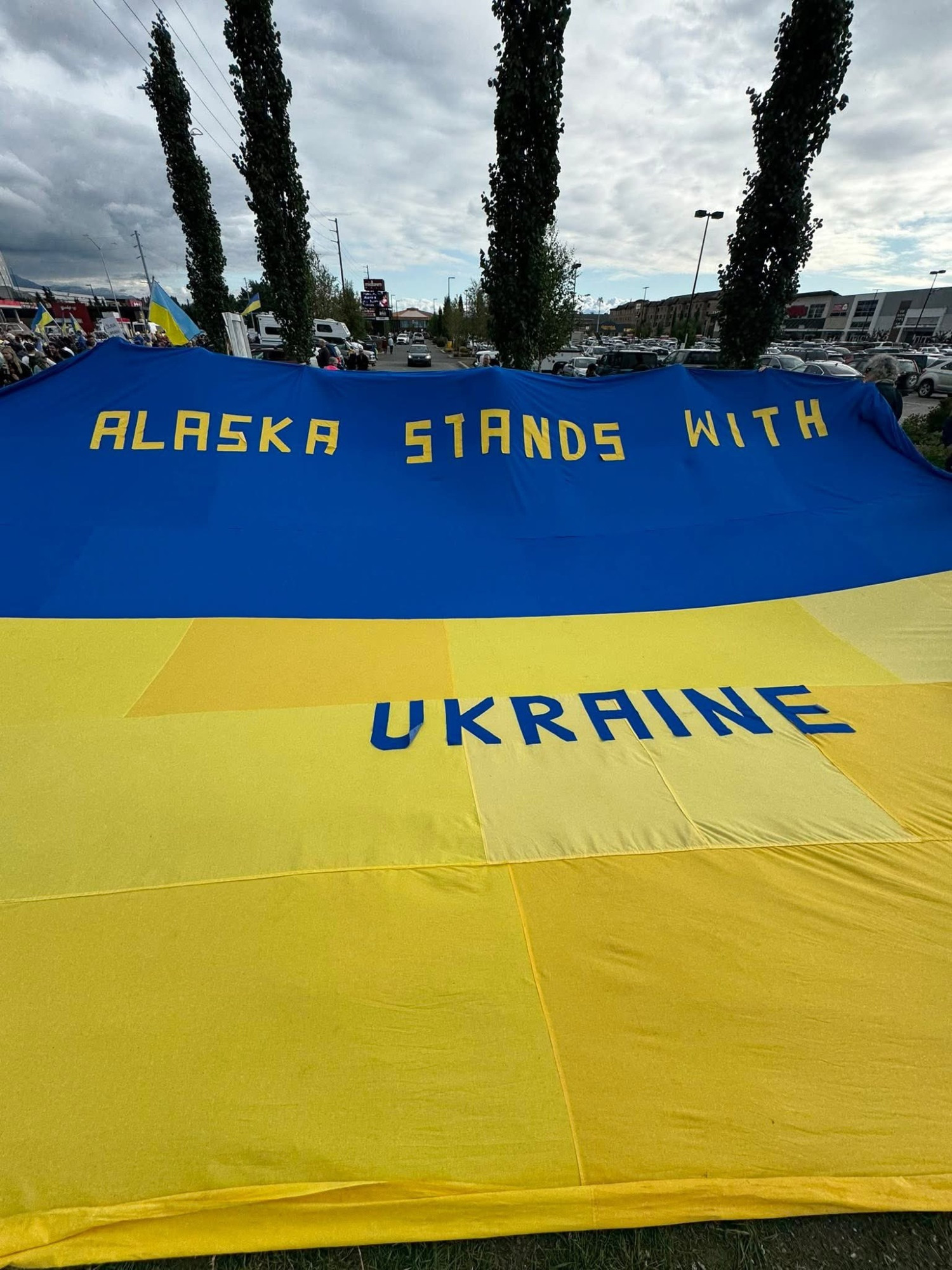 A large Ukraine flag featuring blue and yellow colors displays the words "Alaska Stands With Ukraine." Trees and a cloudy sky are visible in the background.
