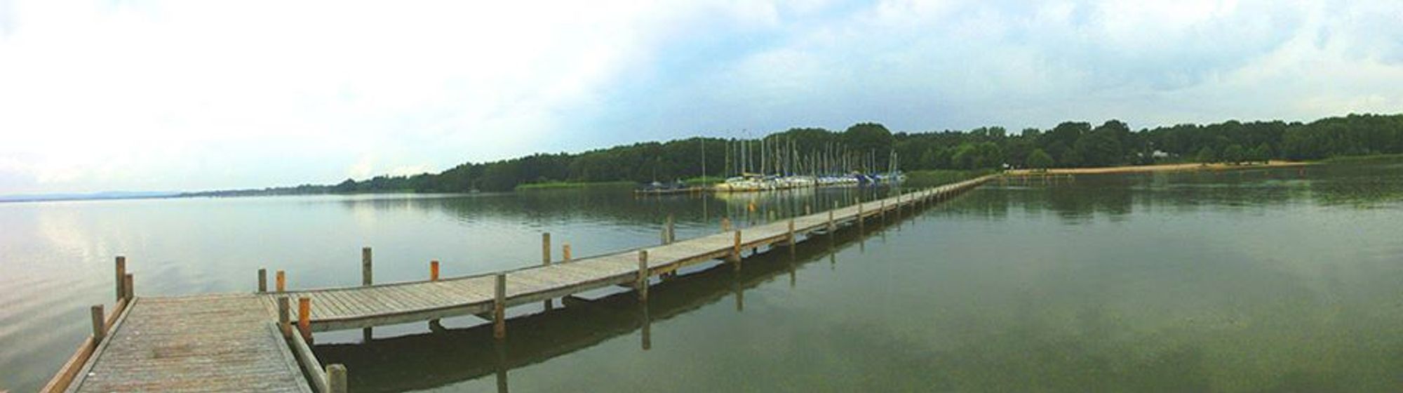 Panoramaaufnahme eines Bootstegs am Steinhuder Meer vom Wasser aus mit Blick auf das Ufer. Das Wasser ist ruhig, der Steg spiegelt sich darin. Am Ufer ist auf der linken Seite ein Bootssteg mit kleinen Segelbooten zu sehen, auf der rechten Seite des Stegs ein Sandstrand. Im Hintergrund große Laubbäume.