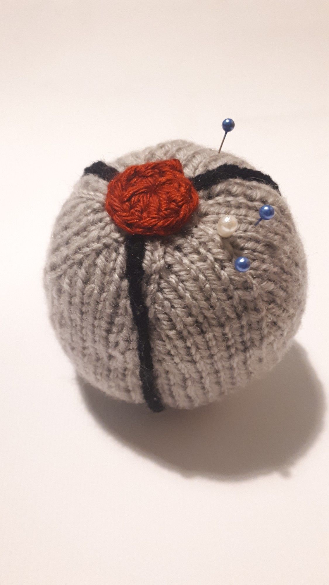 A grey spherical posthuman known as the gravital from All Tomorrows, made into a knitted ball. It has the characteristic seams and red eye. It's pierced by sewing pins with colourful heads. The entire object is set against a matte white backdrop.