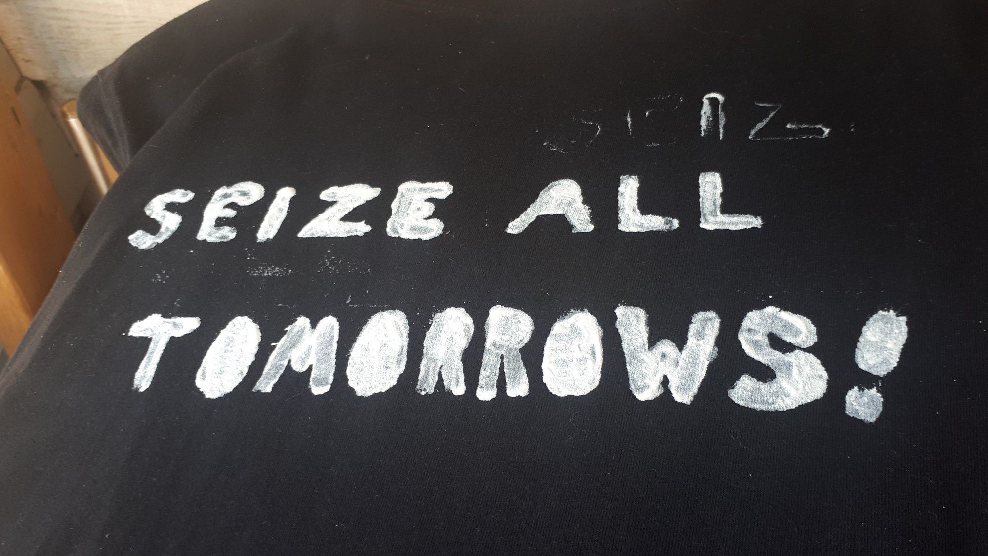 White paint stenciled onto dark fabric that reads "seize all tomorrows!" Another, phantom version of "seize" is above "all" due to the way the paint got under the stencil.