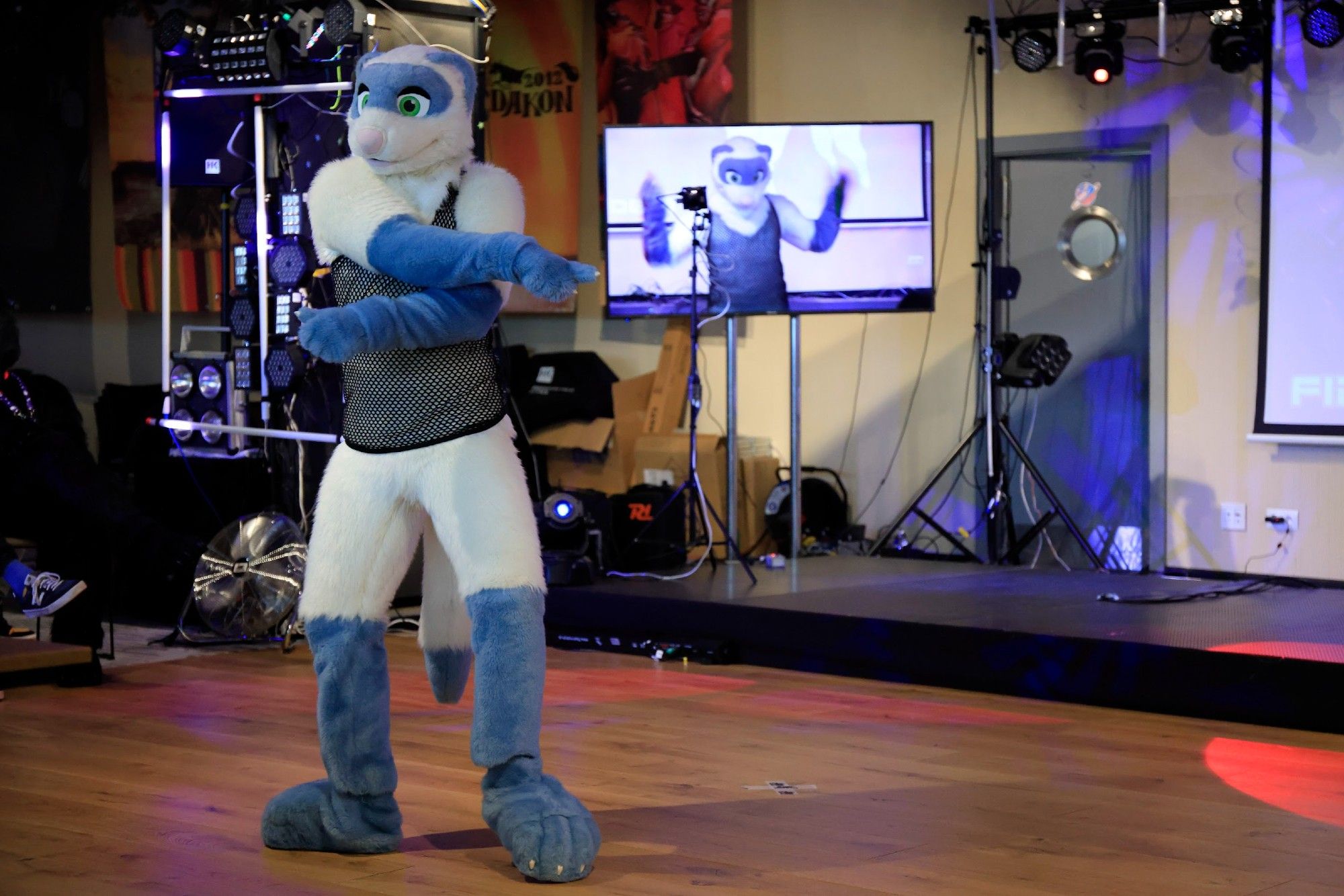 Photo of a white and blue ferret fursuit dancing and pointing at the screen, which shows another picture of the same fursuit doing a different move.