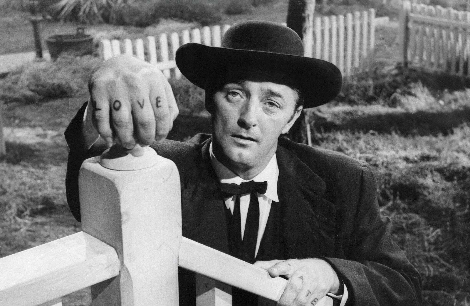 Robert Mitchum as the conman, “Preacher” Harry Powell, holding onto a white stair railing with his hands. The words “LOVE” and “HATE” are tattooed, one letter on each finger of each hand.