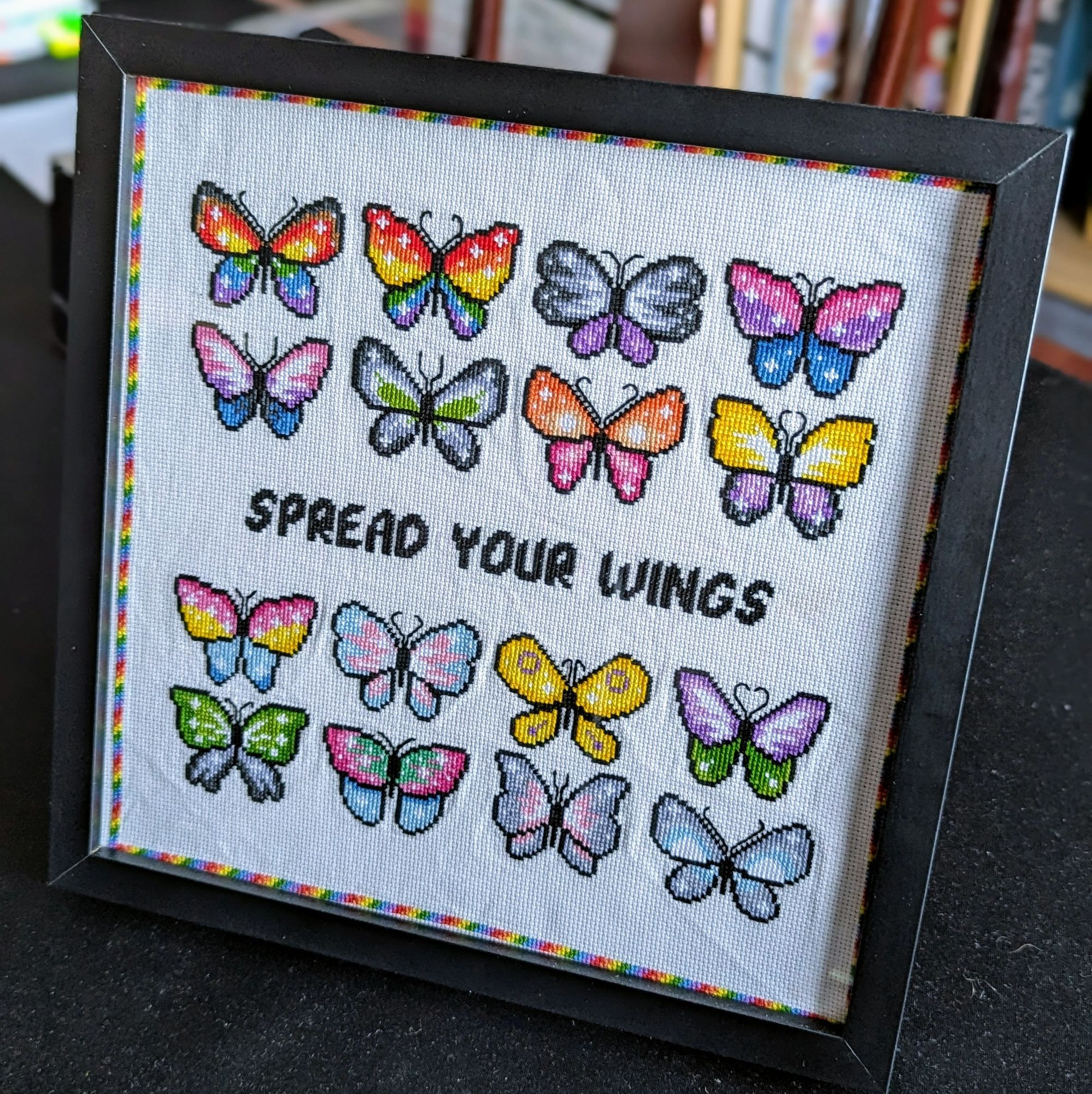 A completed cross-stitch about 10" square, consisting of 16 butterflies, each in the colours of a different LGBTQ+ flag. Across the middle are the words "Spread Your Wings", and around the edge a narrow border in rainbow colours. 
The whole thing is within a square black frame with a glass front.