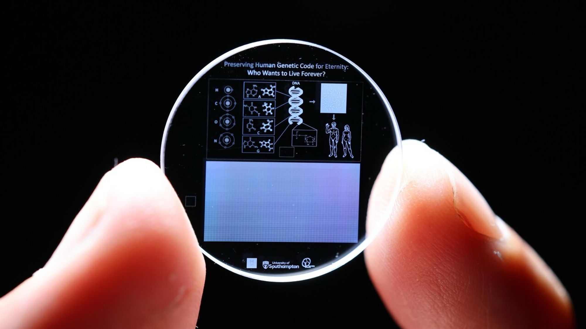 Münzgroße Glasscheibe, gehalten zwischen zwei Fingern, mit winzigen Zeichnungen zur menschlichen DNA, dem im 5D Verfahren gespeicherten menschlichen Genom sowie der Überschrift „Preserving human genetic code for eternity: who wants to live forever?“