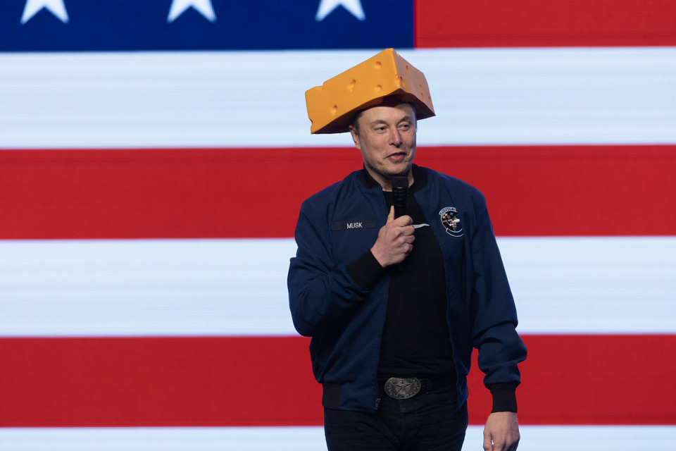 Elon Musk with cheesehead standing with mic in front of American flag