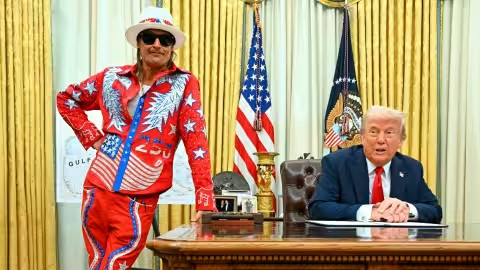 Kid Rock in star-spangled outfit leaning on Oval Office desk with President Trump seated.