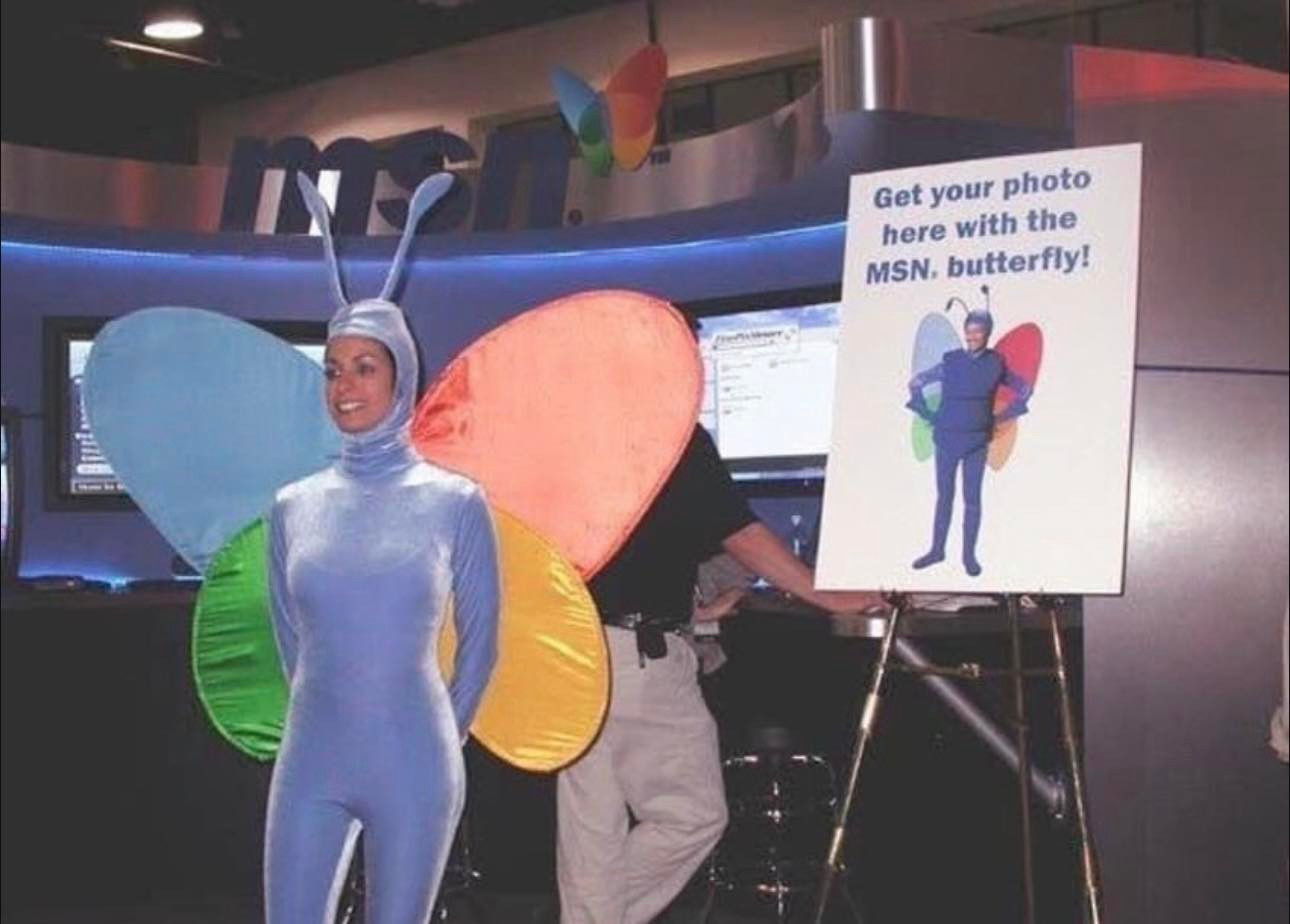 A woman wears a full body costume of a butterfly w/ onlt her face being visible.

Behind her is a sign saying "Get your photo here with the MSN butterfly!"