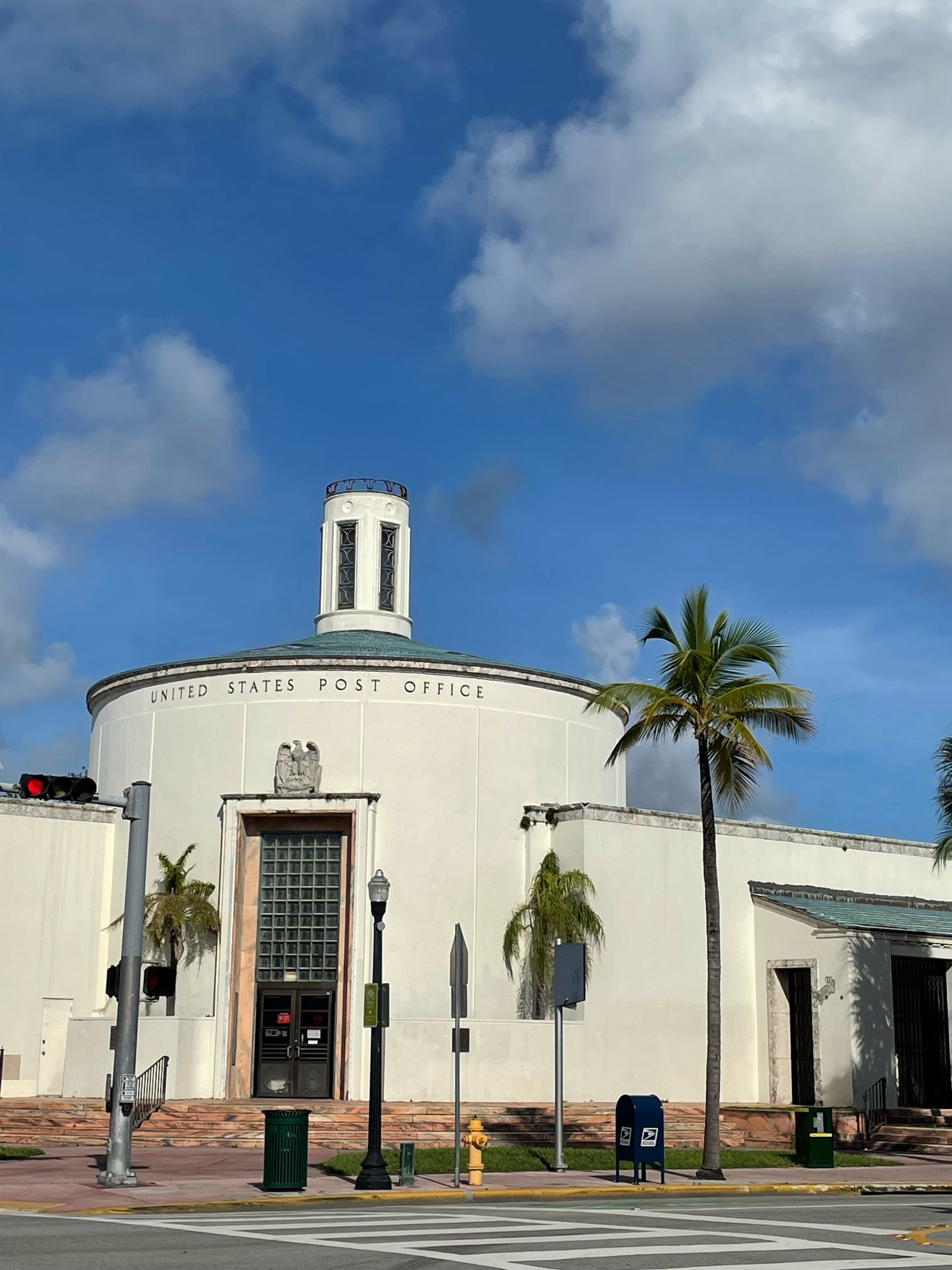 Art Moderne post office building with a tall circular lobby, cone-shaped roof, and a thin tall cupola. Main entrance has “double doors topped by a ten-foot high wall of glass blocks”. Also palm trees.