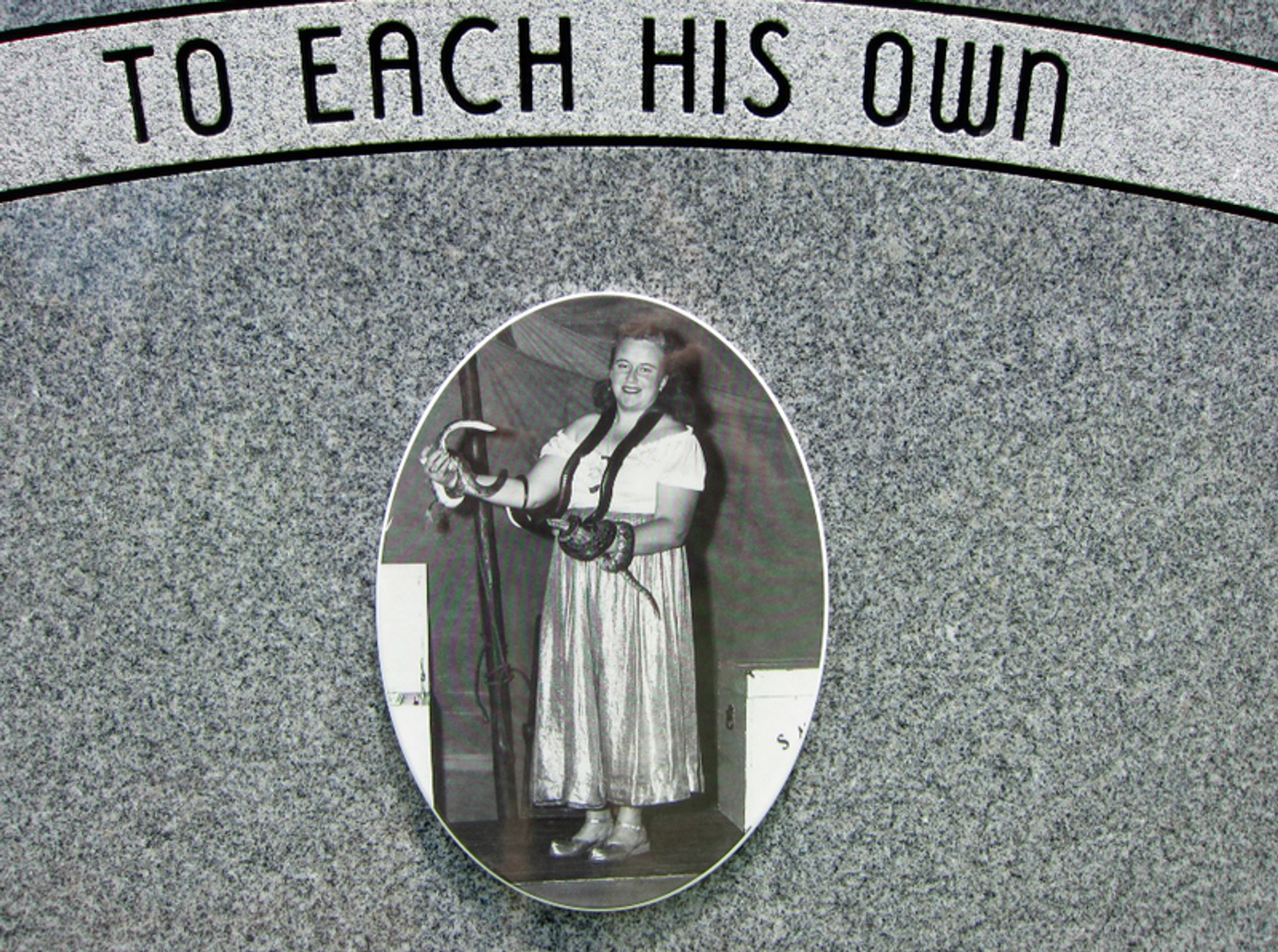 A close-up of a snake-handler's headstone, featuring a photo of a smiling woman in shiny costume dress, holding two snakes. TO EACH HIS OWN is arced over the photo.