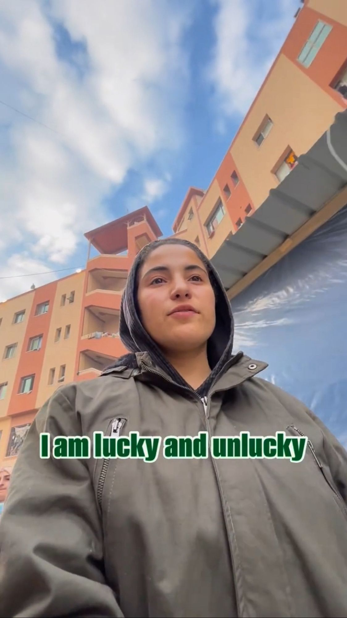 A screenshot of a Bisan Owda video where she is wearing a black hoodie under a green raincoat and walking in front of a tan apartment building and a smaller building with a blue plastic tarp on the side of it. The text on the screen is dark green with white bordering and reads "I am lucky and unlucky".